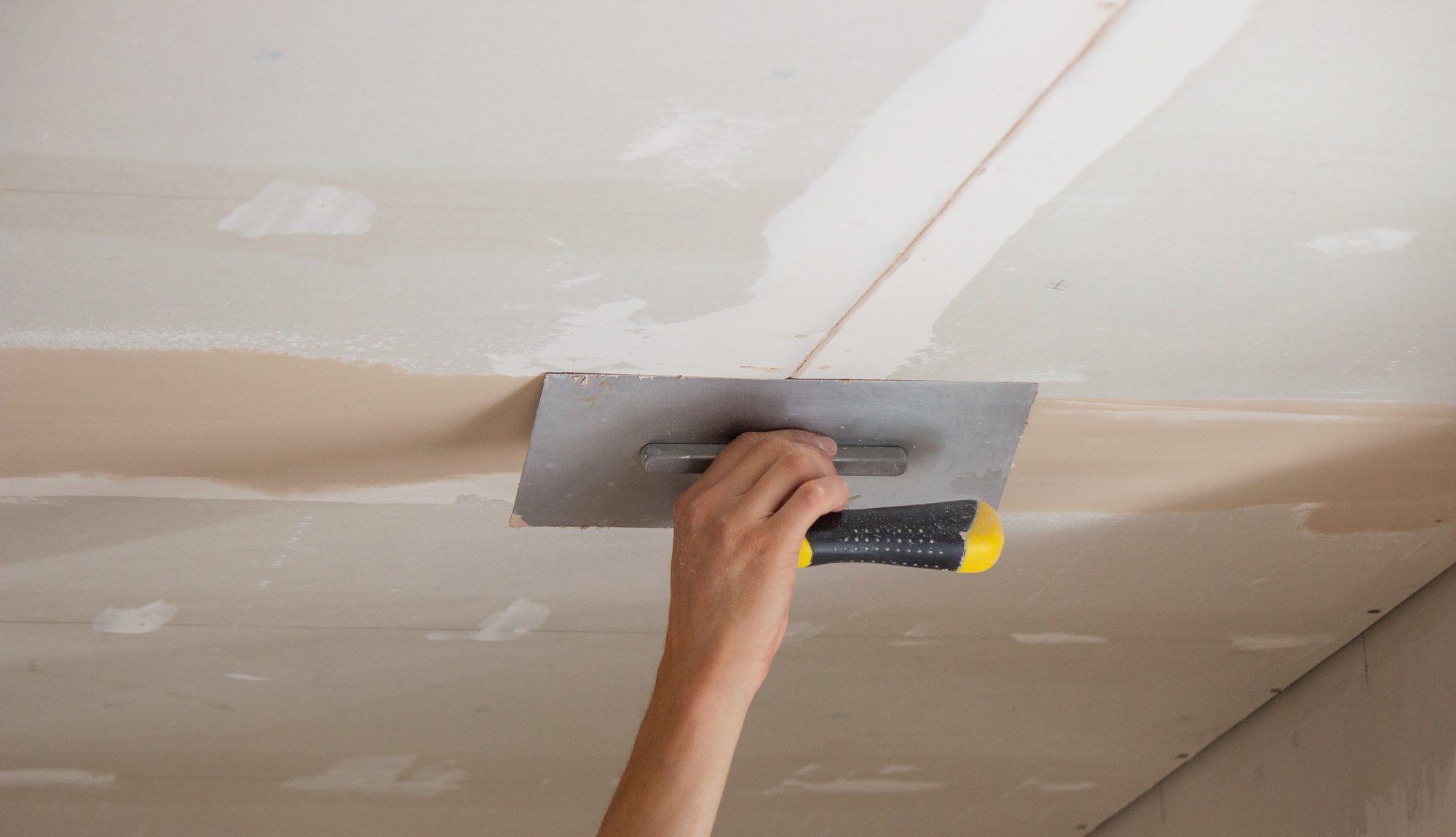 The image shows a person's arm and hand holding a drywall taping knife applying joint compound (also known as mud) to a seam between sheets of drywall on a ceiling. You can see that the compound is being smoothed out, which is part of the process of finishing drywall to create a smooth surface before painting or applying texture. The surrounding area has multiple drywall panels with visible seams and screws that have been partially covered with joint compound that hasn't been smoothed out yet.