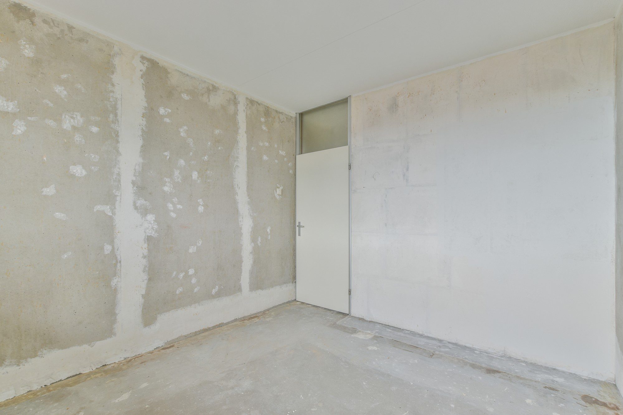 The image shows an empty room with unfinished walls. The walls appear to be in the process of being prepared for painting or re-plastering, as evidenced by the patches where old paint or plaster has been removed, revealing the bare concrete beneath. There's white residue on the floor, which is likely from the wall preparation work. The room has a plain white door that is slightly open, and the ceiling is finished and painted white. It appears to be an interior room in need of renovation or decoration.