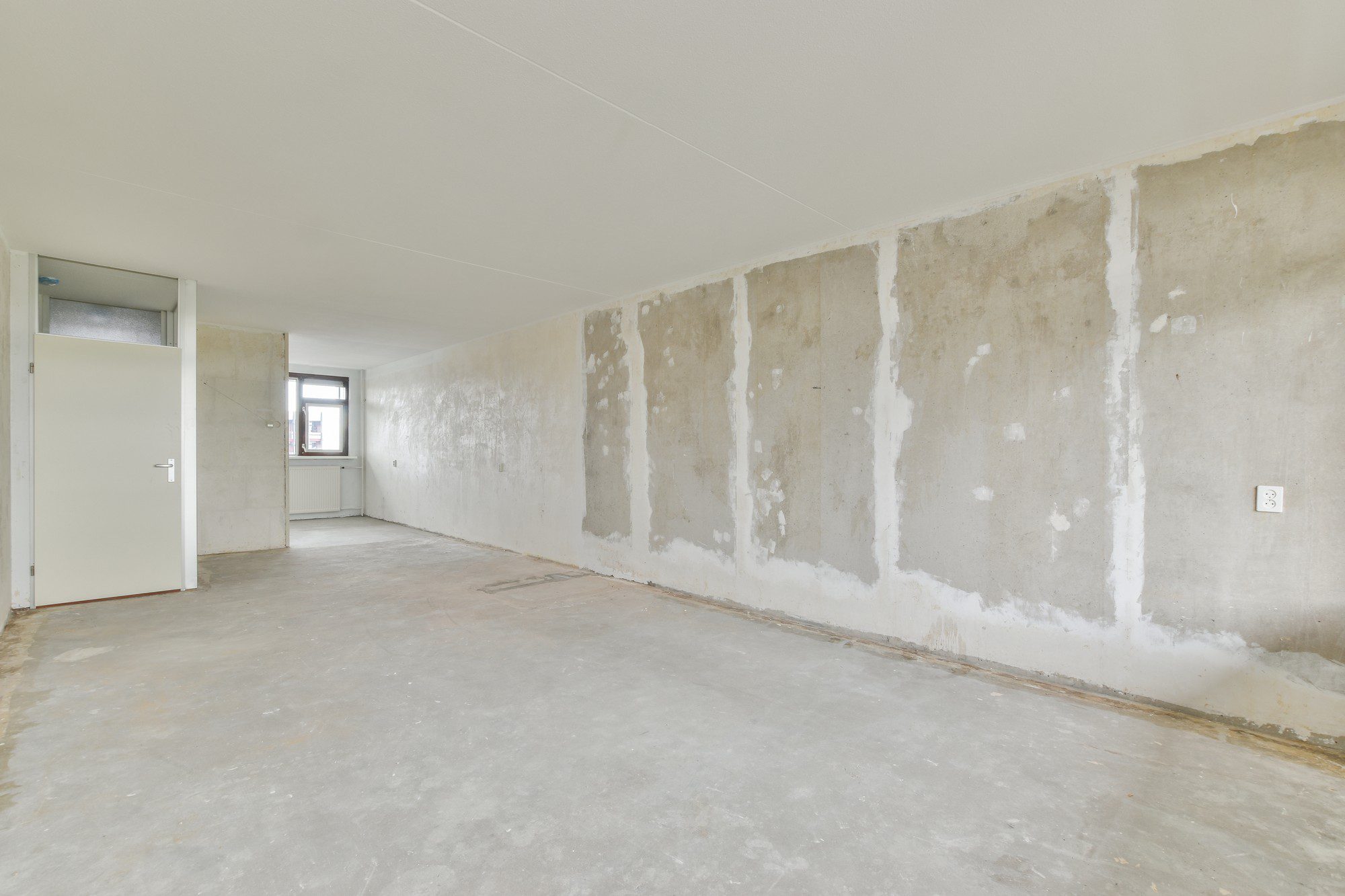 This image shows an empty room that appears to be under renovation or in need of refurbishing. The room has a large, bare concrete wall with patches where possibly previous fixtures or fittings have been removed and the holes have been filled in. The floor seems to be made of concrete as well, and there are no carpets, tiles, or other flooring materials installed.At the far end of the room, there's a window letting in natural light, and on the left, there's an open doorway leading to another room or a hallway. On the left-hand side, a door is partially visible, which looks like it leads to a smaller space, possibly a closet or a bathroom, given the frosted glass part of that door. On the right-hand side of the concrete wall, there's a power outlet. The ceiling appears to be finished and painted white, contrasting with the unfinished state of the walls and the floor.