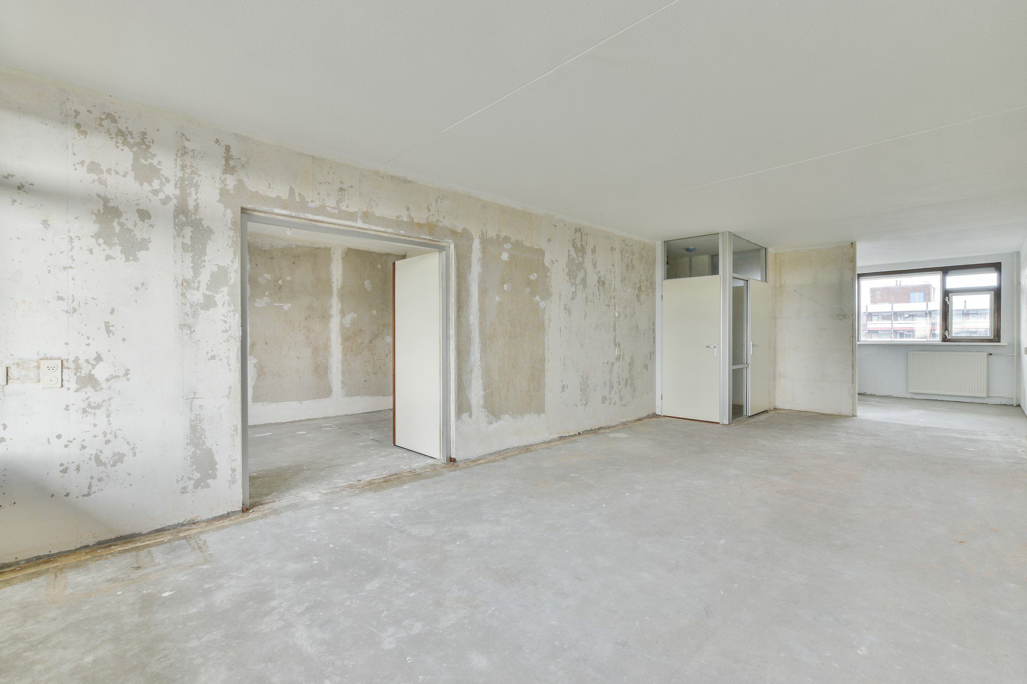 This image shows a vacant interior space that appears to be undergoing renovation or repair. The walls are unfinished with visible patches of peeling paint or plasterwork, suggesting that they might have been stripped in preparation for refinishing. There are no floor coverings, leaving a bare concrete floor. There's an open doorway without a door leading to another room and a white door that appears to lead to a closet or small secondary space. The ceiling is mostly plain with a simple light fixture, and there is a radiator beneath a window in the right corner, indicating that this is likely a residential or office space. The windows allow natural light to enter the room. The general condition of the room indicates that it is a work-in-progress, likely in the middle of renovation.