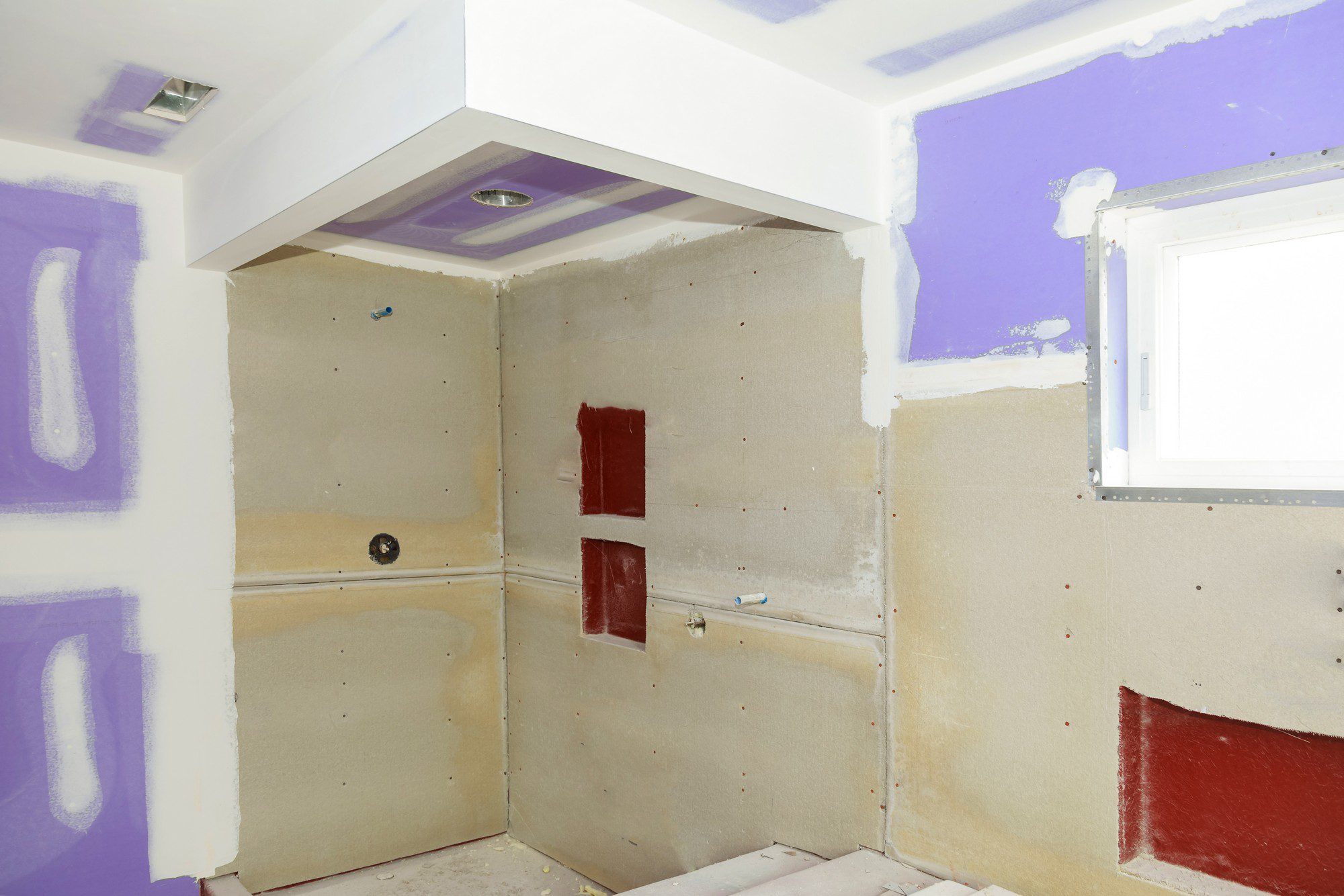 This image shows a room under construction or renovation. The walls are unfinished with visible drywall panels that are not yet painted. Some of the boards are also taped and mudded at the seams, which is a step before sanding and painting. There are patches of purple paint on one wall as if someone has been testing paint colours. On the lower part of the wall, there is a section with red waterproofing membrane applied, which suggests that this area might be prepared for tiling, commonly used in wet areas such as bathrooms or kitchens.

On the ceiling, there are two recessed lighting fixtures already installed. In the wall, there appear to be plumbing and electrical outlets that are roughed in, including a place for what might be a tap or a showerhead, and a small window on the right side of the room. The floor is covered with protective material suggesting that construction work is ongoing. Overall, the image depicts a room that is in the process of being finished.