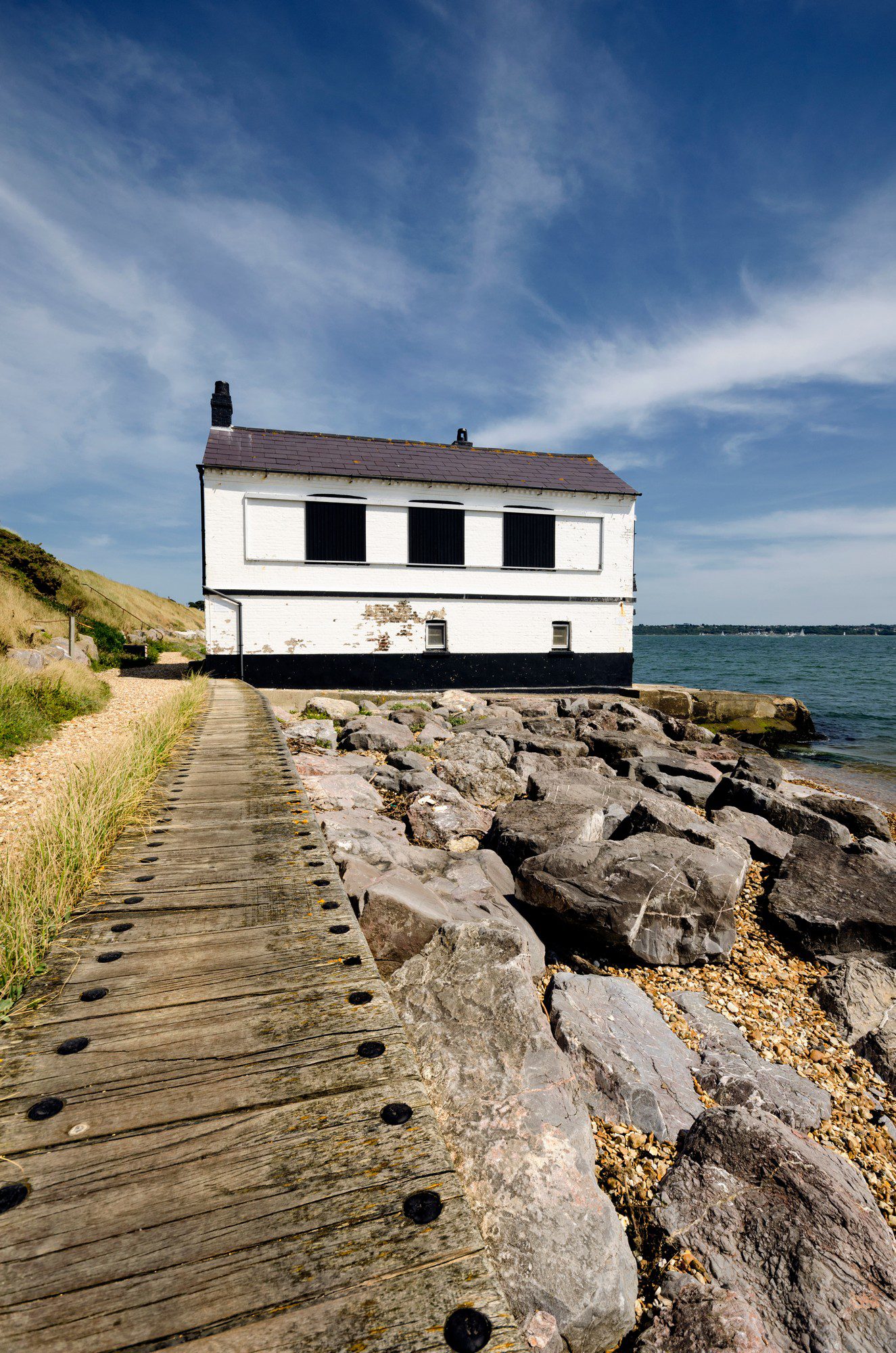 The image shows a picturesque scene featuring a two-story white house with black shutters located right by a body of water. A wooden walkway bordered by large rocks leads up to the house. It seems to be a coastline with some pebbles and smaller stones scattered between the rocks. The landscape includes a grassy hill beside the house, indicating that the dwelling may be somewhat isolated or in a rural coastal area. The weather appears to be sunny, with some clouds scattered across the blue sky, creating a tranquil and scenic atmosphere.