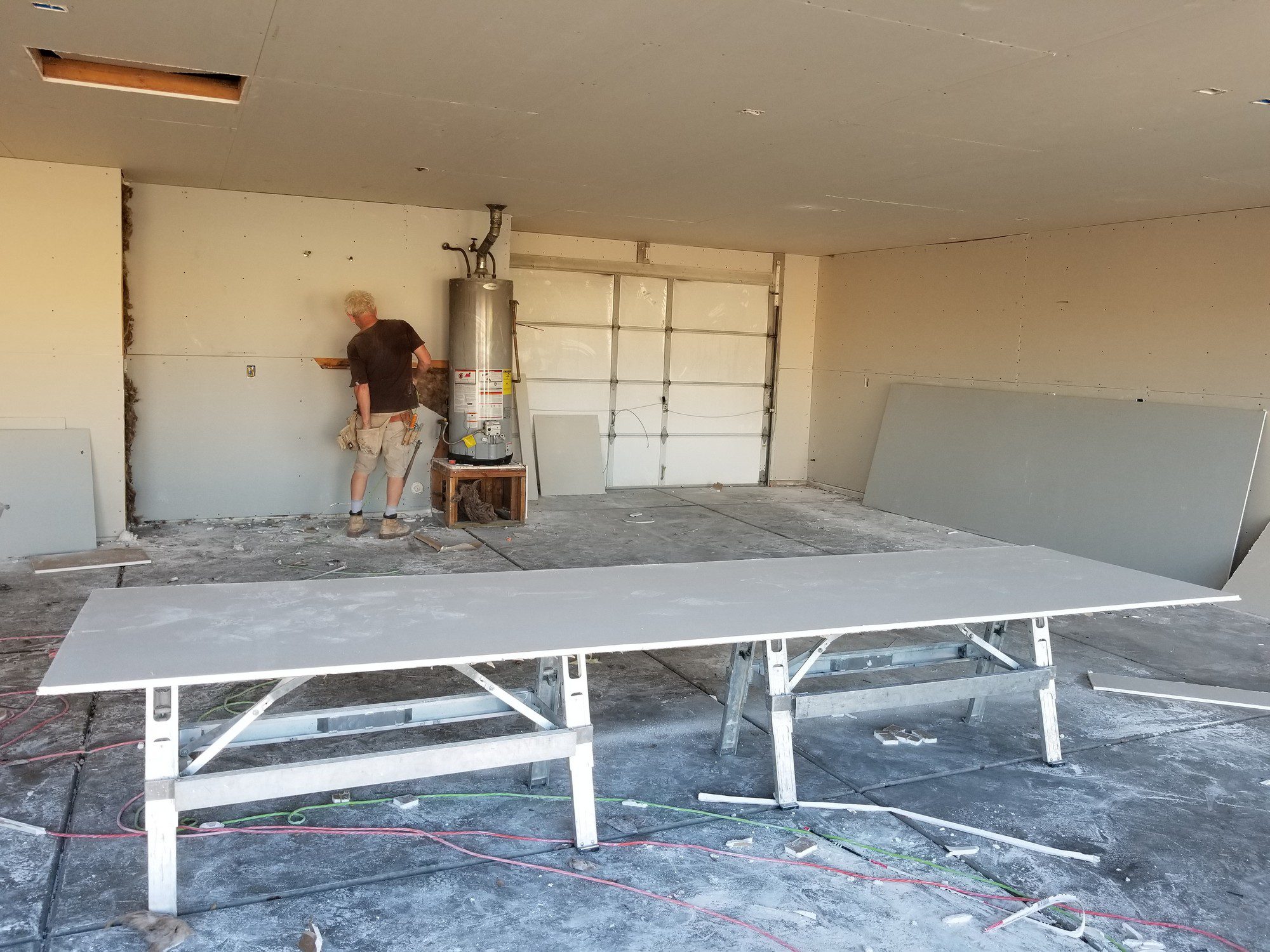 The image shows a person standing in a garage or workshop environment that is undergoing some kind of renovation or construction work. There are large sheets of drywall laid horizontally on sawhorses in the foreground and one leaning against the wall in the back. To the right, a tall water heater is visible. The person appears to be working on something against the wall, possibly related to electrical or construction tasks. The floor is covered with dust and debris, suggesting recent work. The walls are partially covered with drywall, and there is an open space in the ceiling where a panel is missing, exposing the wooden beams. Electrical cords are strewn across the floor, and the overall look indicates an active work site.