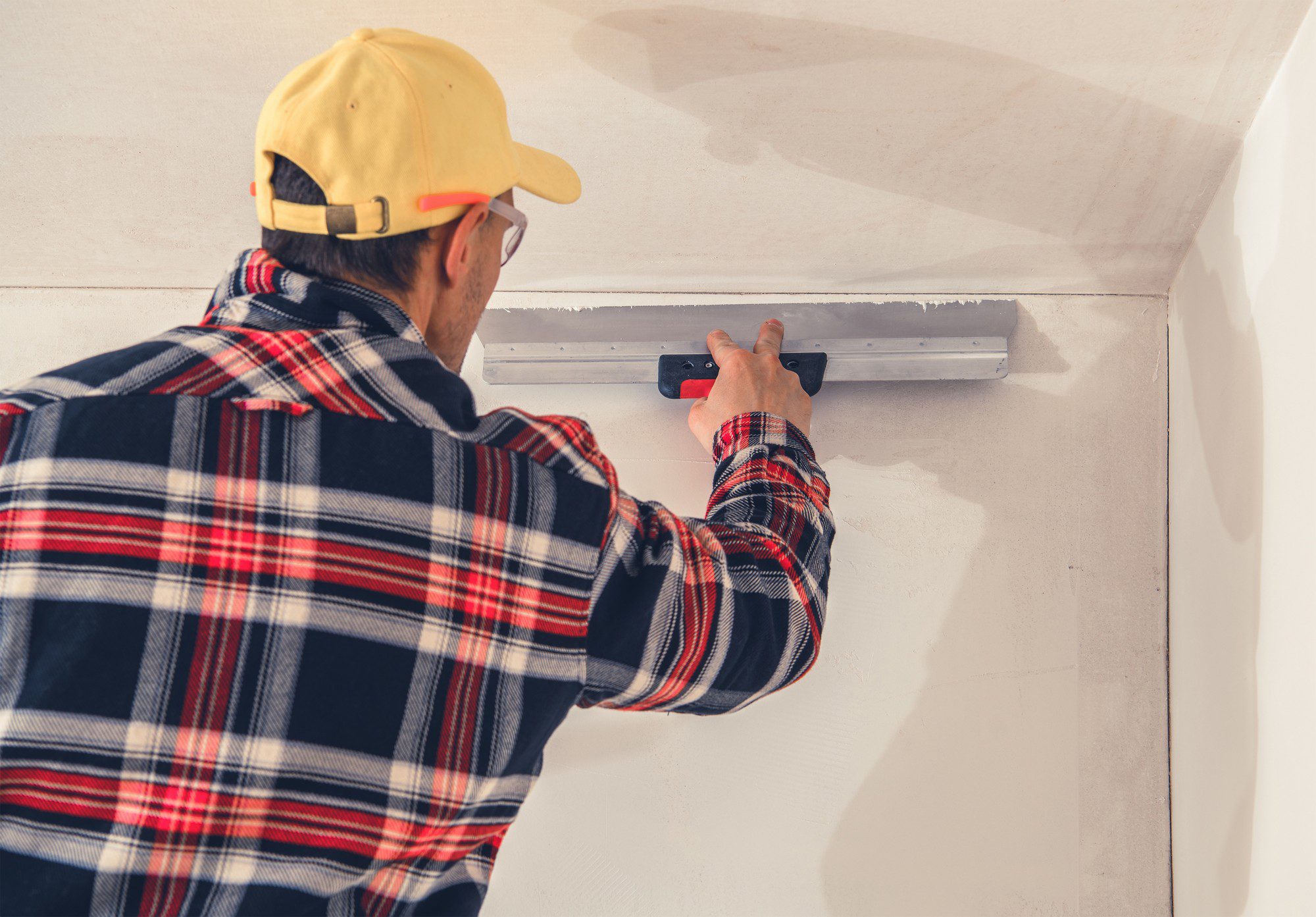 The image shows a person working on a wall. They are wearing a yellow cap, glasses, and a plaid shirt. The individual appears to be using a plastering trowel to apply or smooth plaster or a similar material on the wall, as part of a finishing process in construction or renovation work. The wall has a smooth, off-white surface, and we can see an area where the material has been freshly applied above the trowel. It appears to be an indoor setting, possibly part of a residential or commercial building. The person is focused on their task, ensuring the surface is even and flat.