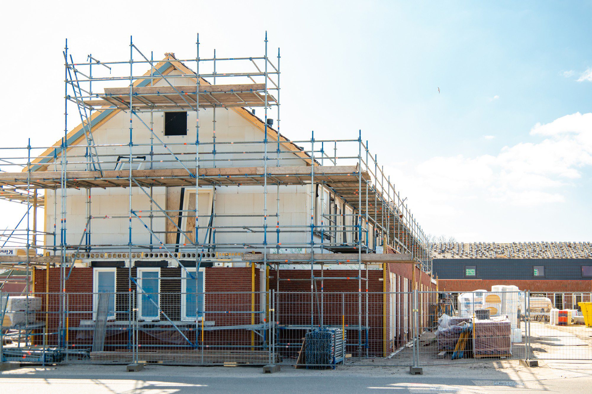 This image shows a construction site for a residential building. There's scaffolding erected all around the partially constructed house, indicating work on the exterior, likely for tasks such as bricklaying, plastering, or installing windows. The building appears to have two stories and an attic or loft space, as suggested by the roof framing. Temporary fencing is in place for safety and security, and there are various construction materials and portable toilets on the site. In the background, there are other buildings with solar panels on their roofs, and a clear sky above indicates good weather, which is favorable for construction activities.