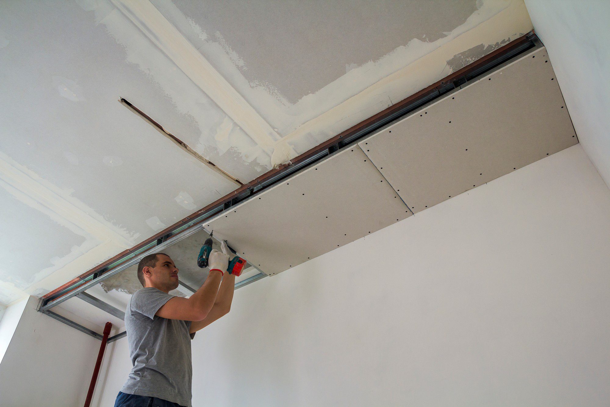 The image shows a person installing drywall or gypsum board on a ceiling. He is using a cordless drill to fasten the drywall panels to what appears to be a metal framework. The ceiling has several panels already in place, but some areas still show the bare frameworks and joint compound used for taping and bedding the drywall seams. It appears to be an indoor construction or renovation scene. The person is focused on the work and is wearing casual clothing, indicative that it may be a do-it-yourself project or a professional working in a casual environment. There's also a red-coloured tool or support structure beneath him, which might be used for holding materials or as a safety feature.