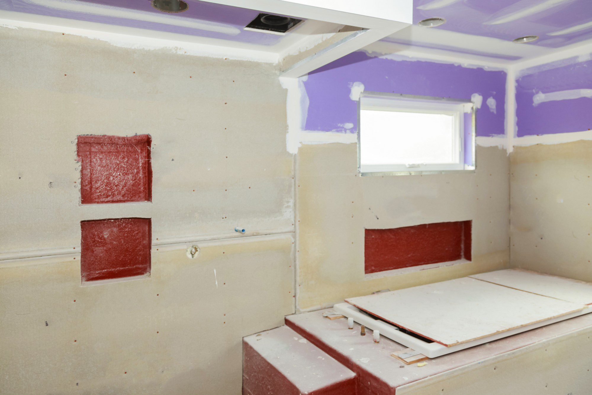 This is an image of a room undergoing renovation or construction. The walls are stripped down to the drywall, and there are patches of joint compound visible where the seams between the drywall panels are being covered. There are several red rectangles on the wall, which could be painted patches or an underlying material, but it's not clear what their purpose is without additional context.

The ceiling is painted purple, and there's a white ceiling cutout, likely for an air vent or some other fixture, with two recessed lighting fixtures installed. The window on the right side of the room is small, with a white frame, and lets in natural light. Below the window, there appears to be a built-in area with some construction material or part of a fixture lying on it. A blue electrical box can be seen sticking out of the wall; there's no electrical device installed on it yet.

The floor is not visible; however, the structure suggests this could be a bathroom or a similar type of space due to the layout and the presence of what might be part of a built-in vanity or tub structure on the right. The room is clearly still a work in progress, with tools and materials likely just outside the frame.