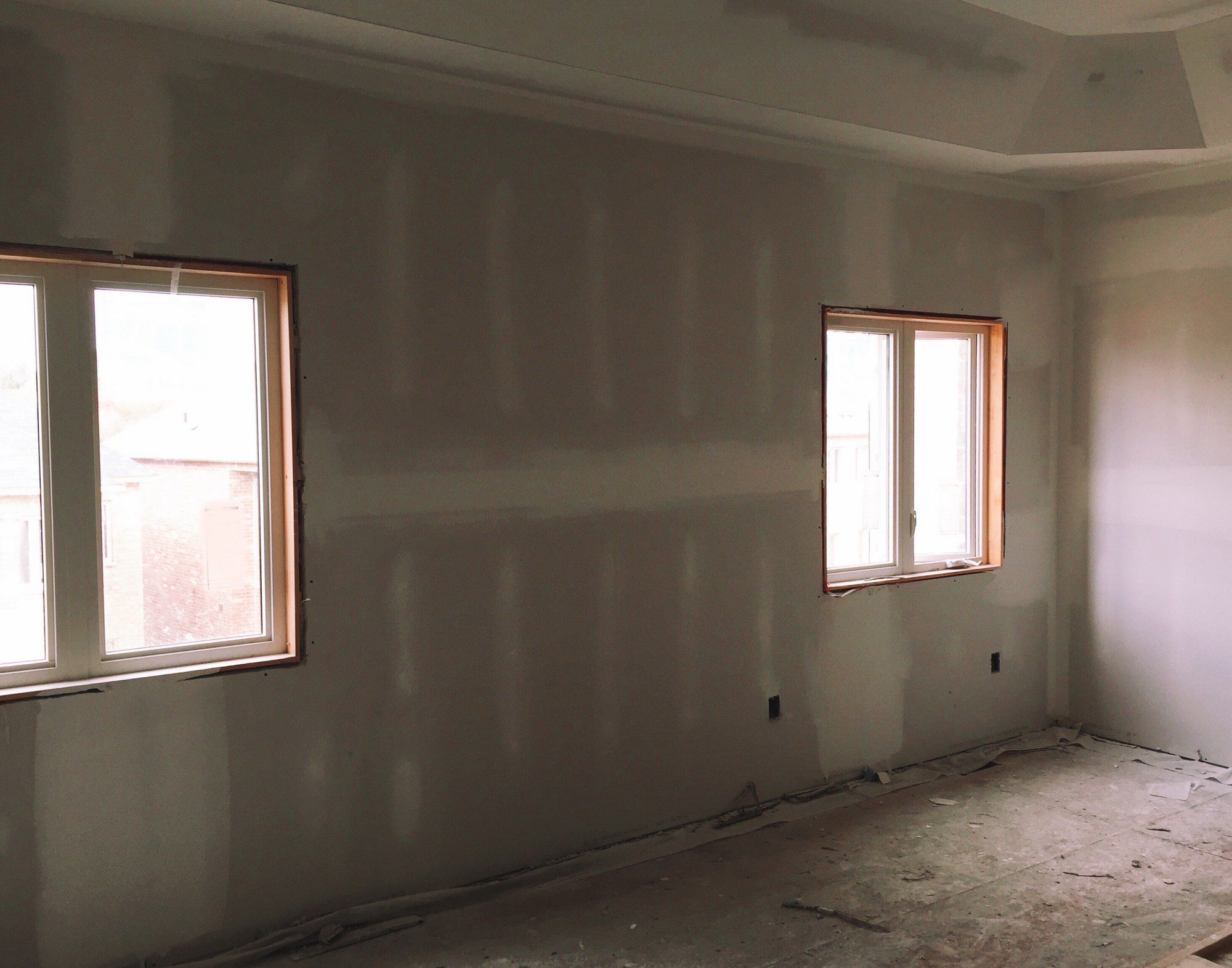 The image shows an interior room that appears to be under construction or renovation. Here are the notable features:1. Two windows allowing natural light into the room, with protective tape around their frames, likely to prevent damage during the construction process.2. Drywall that has been installed on the walls, with the seams and screw holes covered with joint compound, which has yet to be sanded smooth.3. An unfinished floor, which may be a subfloor ready to have flooring material installed over it.4. Electrical outlets have been installed at standard height, indicating readiness for electrical fittings and fixtures.5. The ceiling has a tray or coffered design, adding an architectural element to the room.6. There is no furniture or other fixtures in the room, which is common for a space that is still being worked on.Overall, the room is in an intermediate stage of construction, with further finishing work required before it's completed.