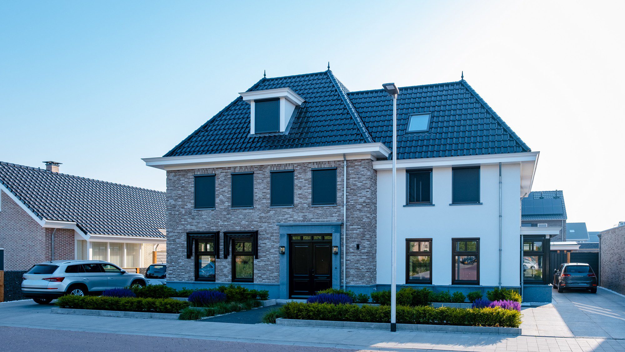 This image shows a modern two-story house with a symmetrical design. The house has a mixture of brickwork and white plastered facades and features a steep gabled roof with dark roof tiles. There are dormer windows on both sides of the roof, enhancing the symmetry. In front of the house, there's a well-maintained garden with flowering plants and shrubs, and the entrance has a dark-coloured front door with a small overhang for shelter.

There's also a paved driveway leading up to a garage on the right side of the house, and two cars are parked — one in the driveway and the other in front of the house. The clear blue sky suggests it might be a sunny day, and the overall appearance is that of a quiet suburban neighborhood.