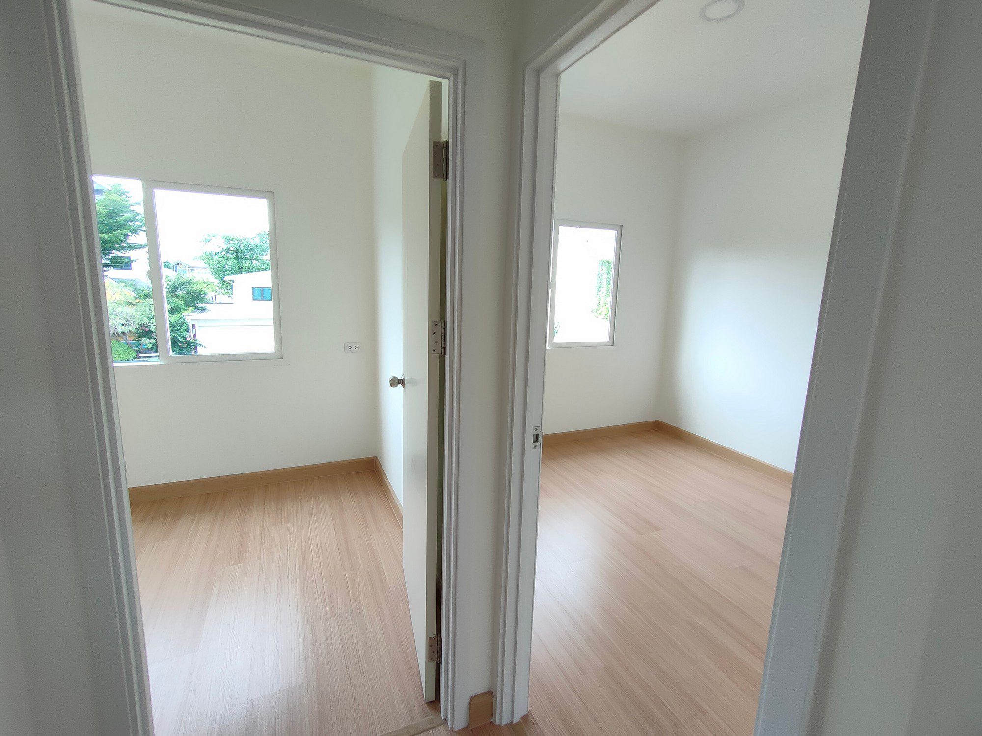 The image shows an empty room with an open doorway leading into it. The walls of the room are painted white, and there is a light wood laminate flooring. Natural light is coming in through two windows, one visible through the doorway and another on the wall to the right. There are no furnishings or decorations in the room, giving it an uninhabited and clean look. The room appears to be ready for occupancy or might have just been renovated.