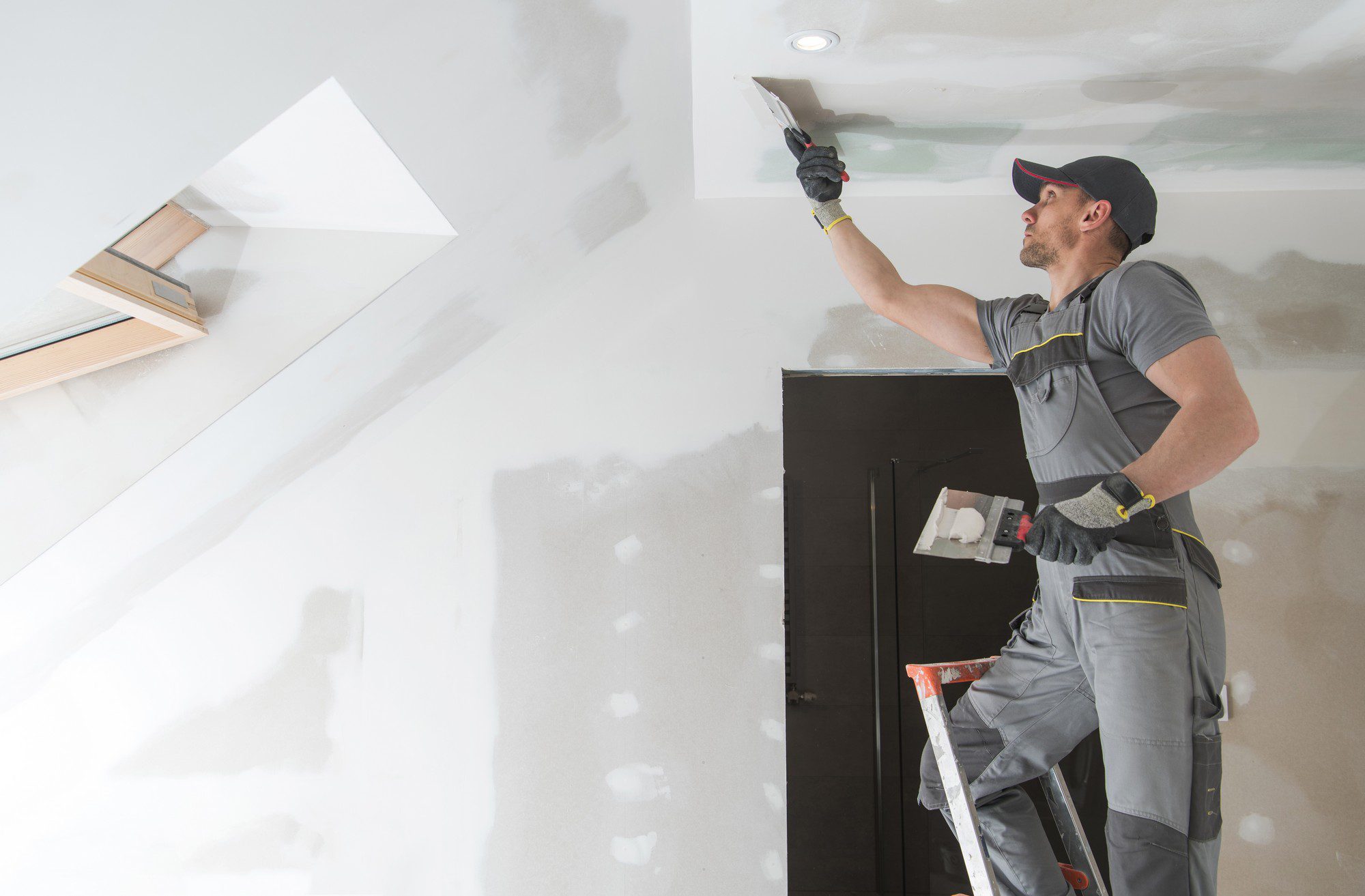 The image shows a person working on a drywall ceiling installation or repair. The person is holding a plastering trowel, applying material to the joints of the ceiling sheets to create a smooth finish. They are standing on a stepladder to reach the ceiling. The worker is wearing a cap, a pair of gloves, and a work outfit with reflective safety stripes. Visible in the surroundings are patches on the ceiling that have not yet been smoothed over, a recessed ceiling light, and a skylight or a loft window. The overall scene is an indoor construction or renovation setting with work in progress.