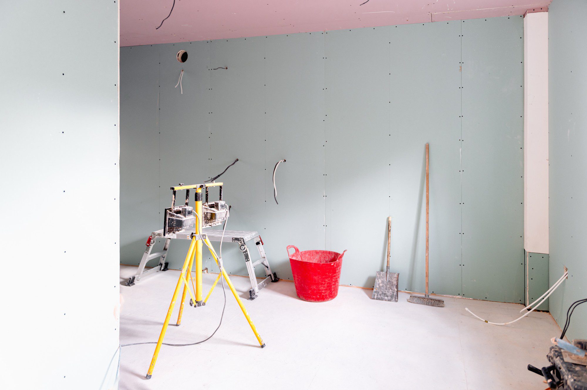 The image shows a room that appears to be under construction or renovation. The walls are partially covered with plasterboard (drywall), and there's visible insulation in the ceiling. Various construction tools and materials can be seen, including:

1. A yellow and silver stepladder in the centre, likely being used to reach higher areas of the walls or ceiling during work.
2. A red bucket on the floor to the right of the ladder, possibly for holding construction materials or waste.
3. An electricity cable reel with a black power cord, resting on the floor near the ladder.
4. Cables hanging loose from the walls and ceiling, likely for future electrical installations.
5. A silver shovel and a push broom leaning against the wall on the right side, which are typically used for clean-up operations on a construction site.
6. Additional plasterboard pieces leaning against the wall, which will probably be used to complete the wall surfaces.
7. The floor is protected by a covering, which might be to prevent damage or staining during the work.

The unfinished state of the room suggests that work is actively ongoing, and the room will undergo further changes as the renovation progresses.