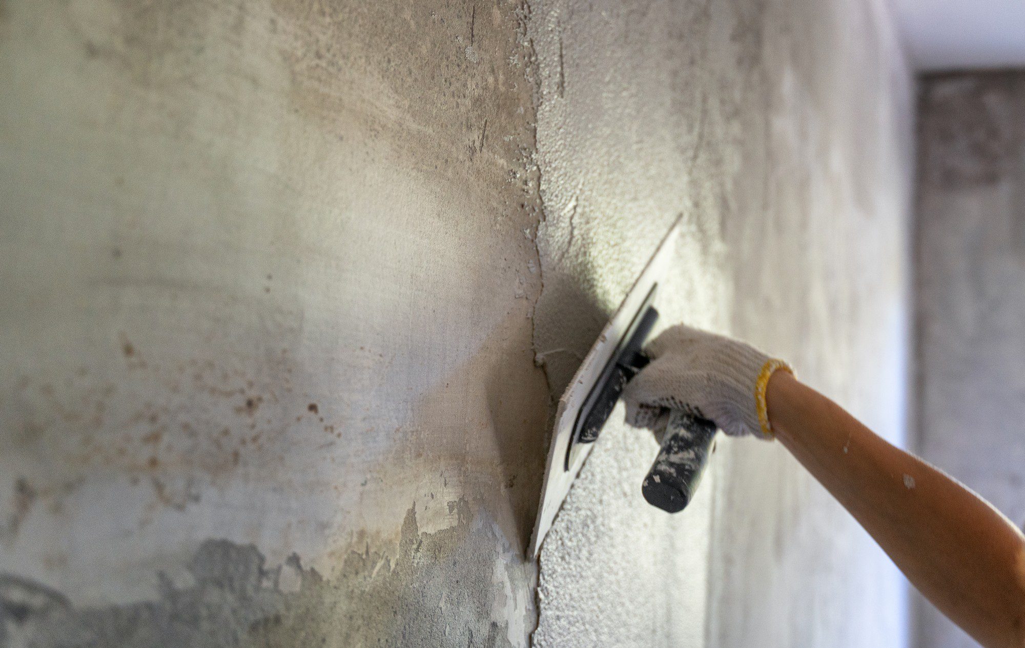 The image shows a person applying plaster or putty on a wall using a trowel. The person's hand is gloved, indicating that they are taking precautions to protect their skin during the work. The work is part of a wall surface preparation process, which could be for painting, wallpapering, or simply to smooth the wall surface. The wall appears to have some uneven patches and possibly some moisture damage or old paint remnants that are being covered or smoothed out.