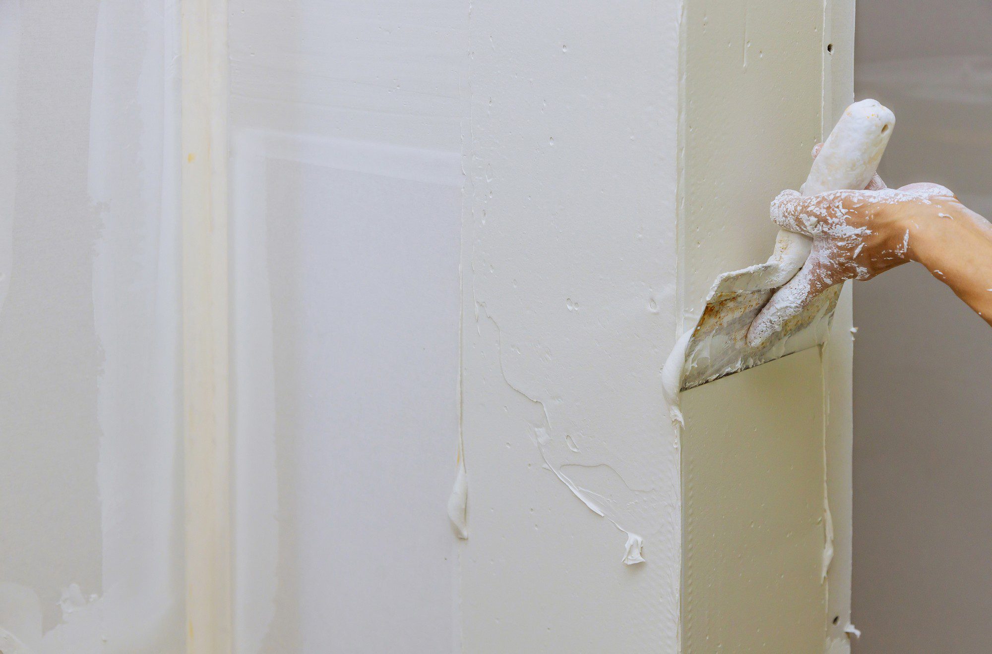 The image shows a close-up view of a partially painted surface, with white paint that appears thick and possibly still wet, given the way it's running down the wall. A person's hand, covered in paint and wearing a painting glove, is holding a paint scraper or putty knife against the door frame or wall corner, actively scraping off excess or dripping paint. The scene suggests a painting project in progress, with attention being given to smoothing out the painted surface or removing excess material to ensure a neat and finished job.