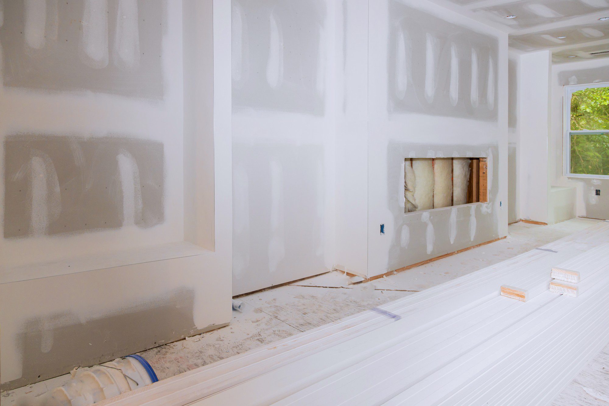 The image shows an interior space undergoing construction or renovation. The walls appear to be freshly drywalled with joint compound applied on the seams and screw indentations, a process known as taping and mudding, which is done before sanding and painting. 

A pile of white primed trim boards is on the floor in the foreground, suggesting that the trim work has yet to be installed. On the right, there's a window letting in natural light. Additionally, there's an opening in the wall with exposed insulation, which could be the site for a fireplace, a vent, or some other feature that is yet to be completed. 

The floor is covered with dust and debris from the construction work, and it looks like protective paper or a drop cloth might be laid down in part to protect the floor. Electrical outlet boxes are visible on the walls, and the room is otherwise empty, indicating that the space is in an intermediate construction stage.