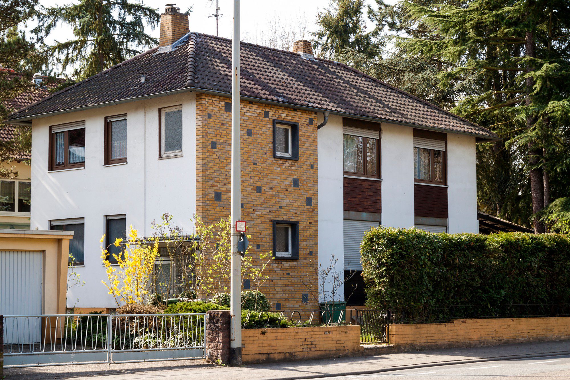 The image shows a two-story residential building. The building features a combination of white rendering and exposed brickwork, with a tiled gable roof. There are several windows with closed shutters on both floors. To the left of the building, there's an attached garage with a light-coloured door. In the foreground, there's a metal fence and gate, partially obscuring a small garden area with shrubs and a budding tree. A street light is also visible on the left side of the image. The setting appears to be in a suburban area, and given the tree in bloom, it might be during the spring season.