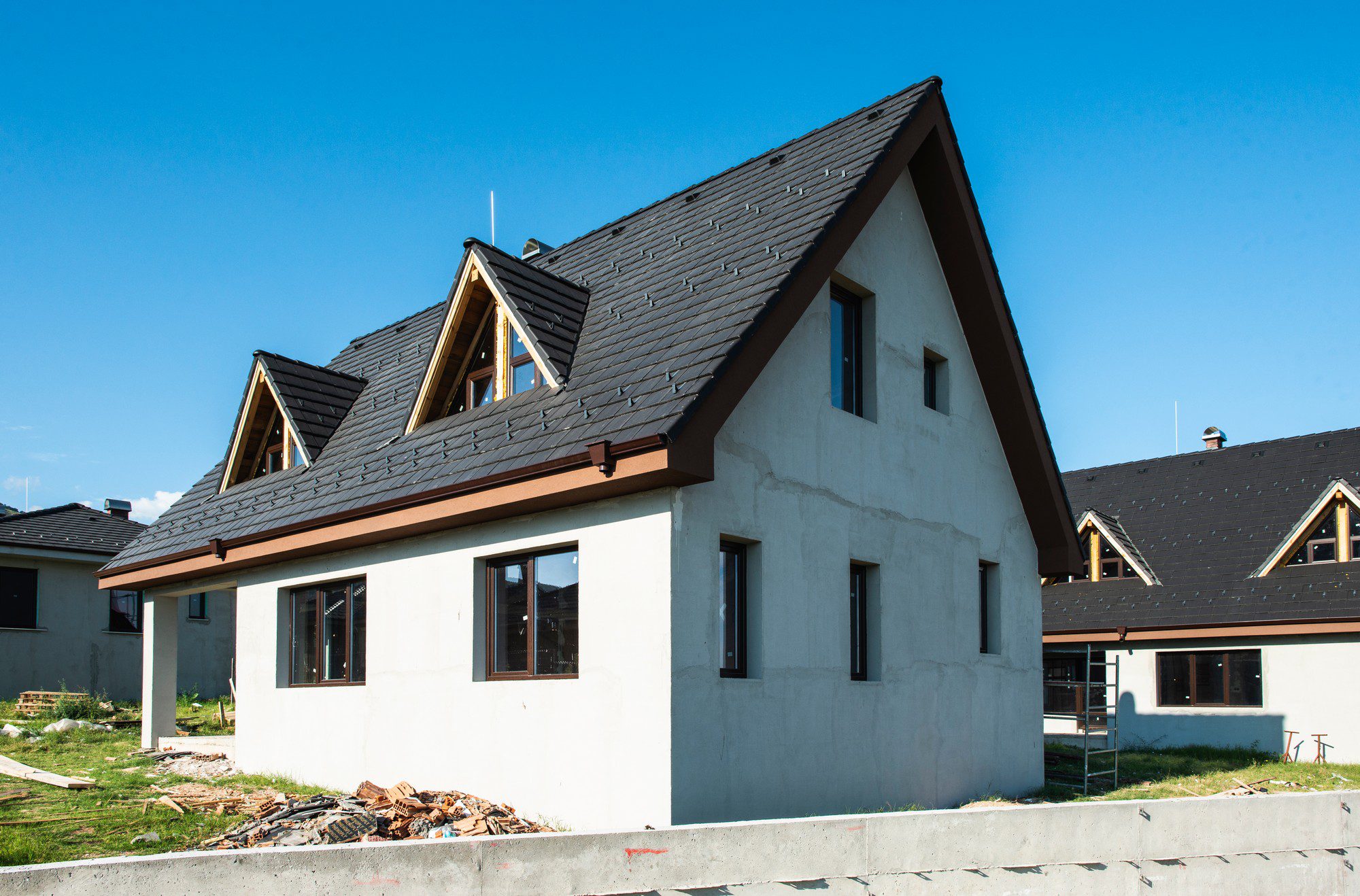 This image shows a residential house under construction. The structure of the house appears complete, with a finished roof, dormer windows, and exterior walls that have been plastered but not yet painted. The house has multiple windows installed, and construction debris is visible around the building site, indicating ongoing work. The photo depicts clear skies, inferring fair weather conditions which are often suitable for construction activities.