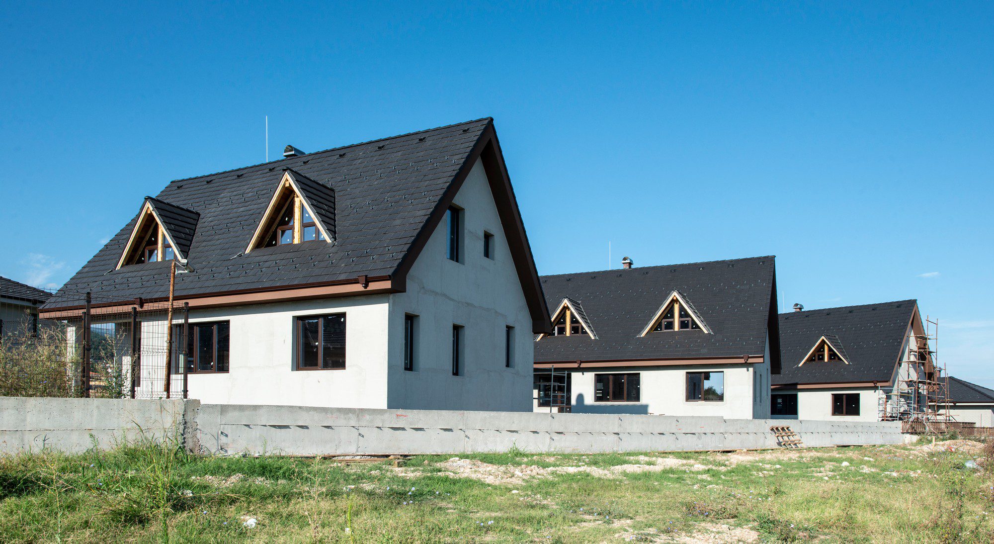 The image shows a partially constructed residential building with two sections that appear nearly identical, featuring a dark-coloured pitched roof with dormer windows. The exterior walls are partially finished with exposed areas, and there's scaffolding visible on the right side, indicating ongoing construction work. The premises are enclosed by a low concrete wall, and the surrounding area is a grassy field with some debris scattered around, typical of a construction site. It's a clear day with a blue sky in the background.