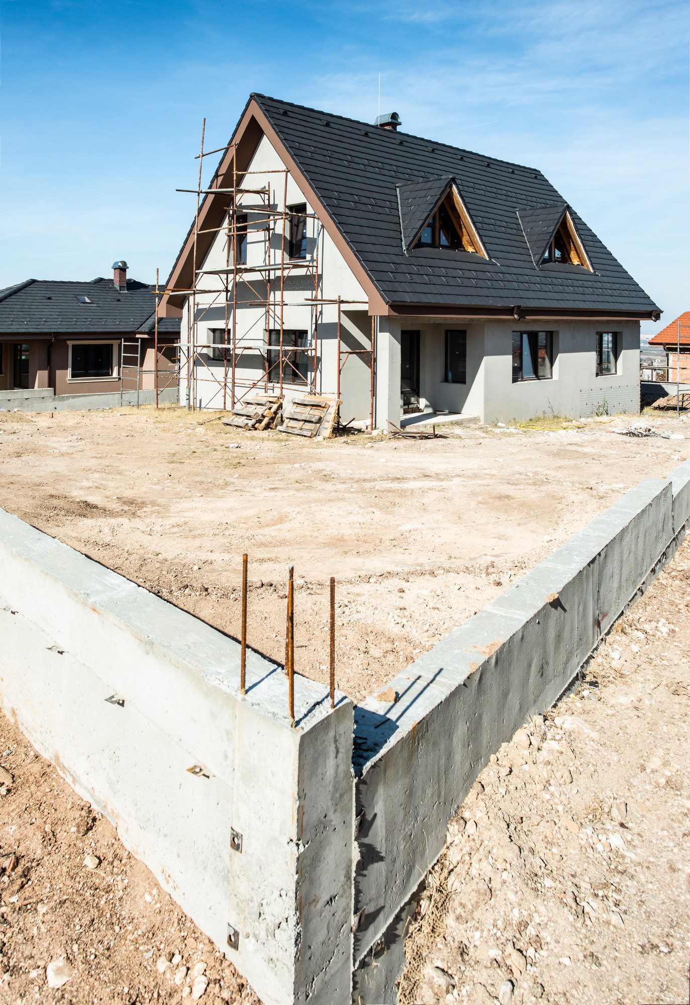 The image shows a residential construction site. In the foreground, you can see a concrete foundation with protruding steel reinforcing bars (rebar), which suggests that more construction might be planned adjacent to this foundation, possibly for another building or an extension.

In the background, there is a two-story house with scaffolding on one side, indicating that work is still in progress. The house has a pitched roof with dark roofing material, and the exterior walls are partially finished. It appears to be a modern design with dormer windows integrating into the roof. The ground around the buildings is bare and looks like it has been cleared or graded recently, likely in preparation for further development or landscaping.

The clear blue sky suggests that the photo was taken on a sunny day, which benefits outdoor construction activities.