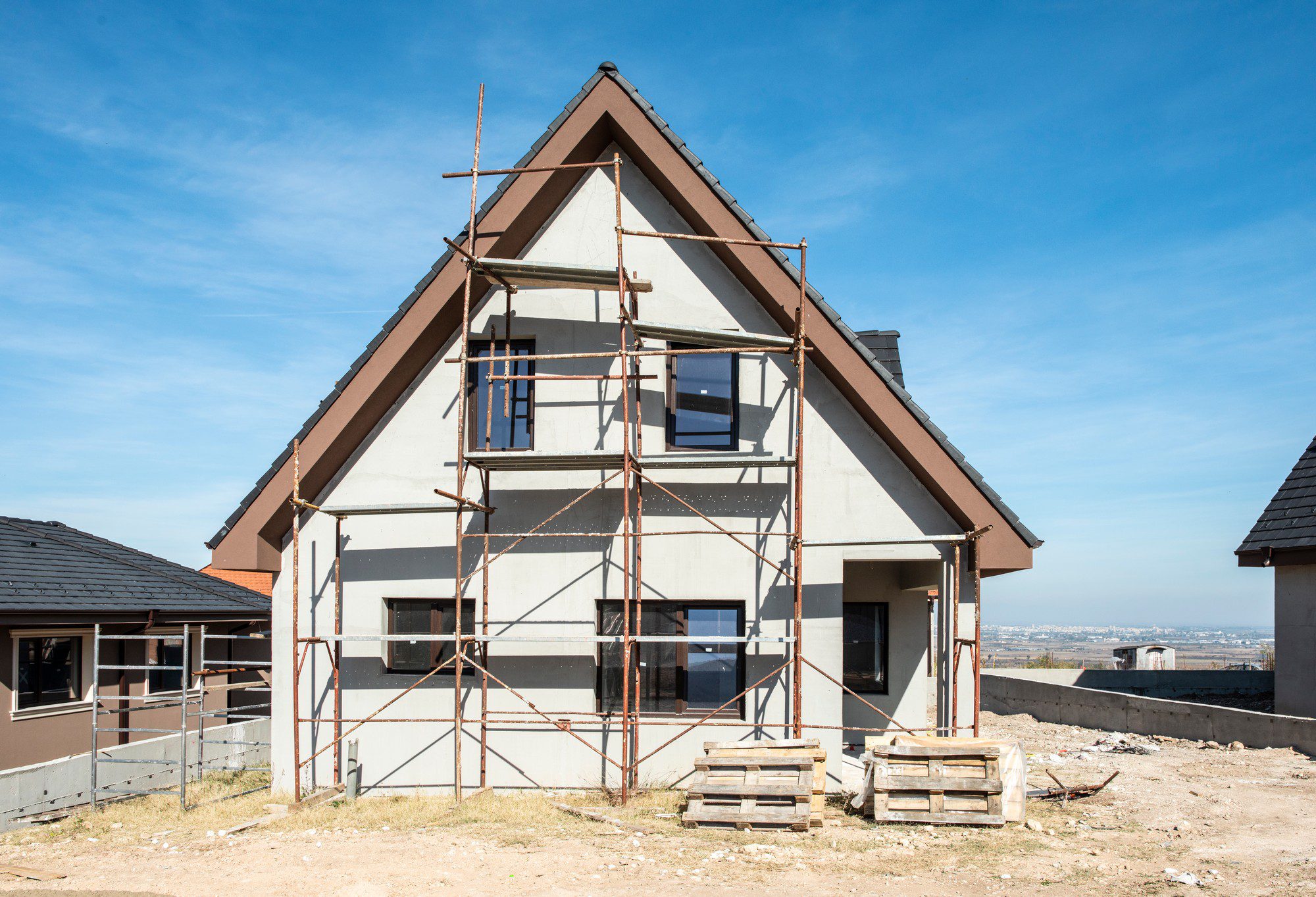 This is an image of a house under construction. The house is at a stage where the basic structure is complete, featuring its roof and window openings. There is scaffolding set up along one side of the house, indicating that work is being done on the exterior, possibly on the facade or the walls. In the foreground, there are pallets and construction debris scattered across the ground, which is common in construction sites. The setting appears to be a developing neighborhood, as there are other houses nearby, and there is a distant view of a city or town in the background, suggesting that this area is on the outskirts of a populated region.