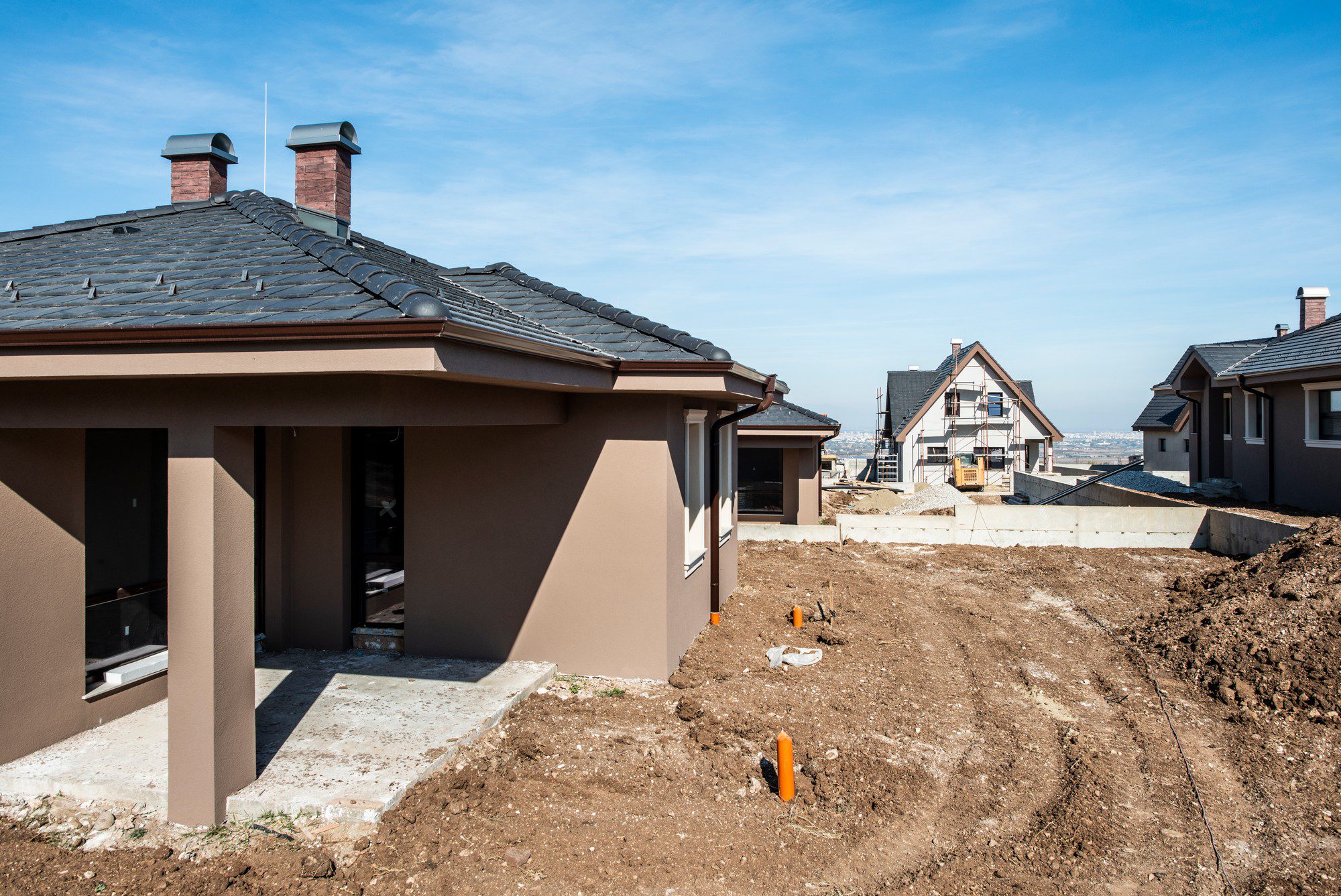 The image shows a residential construction site with newly constructed houses. The foreground features a house with a dark tiled roof and two brick chimneys. There is uncovered soil and construction debris around, suggesting that landscaping and final touches to the property are still pending. You can also see the concrete foundation for what appears to be a patio or porch area directly in front of the house. In the background, another house is visible with scaffolding along its side, indicating ongoing construction or finishing work. The sky is clear, and it looks like the construction site is located in a suburban or rural area, given the open spaces and the distant horizon.