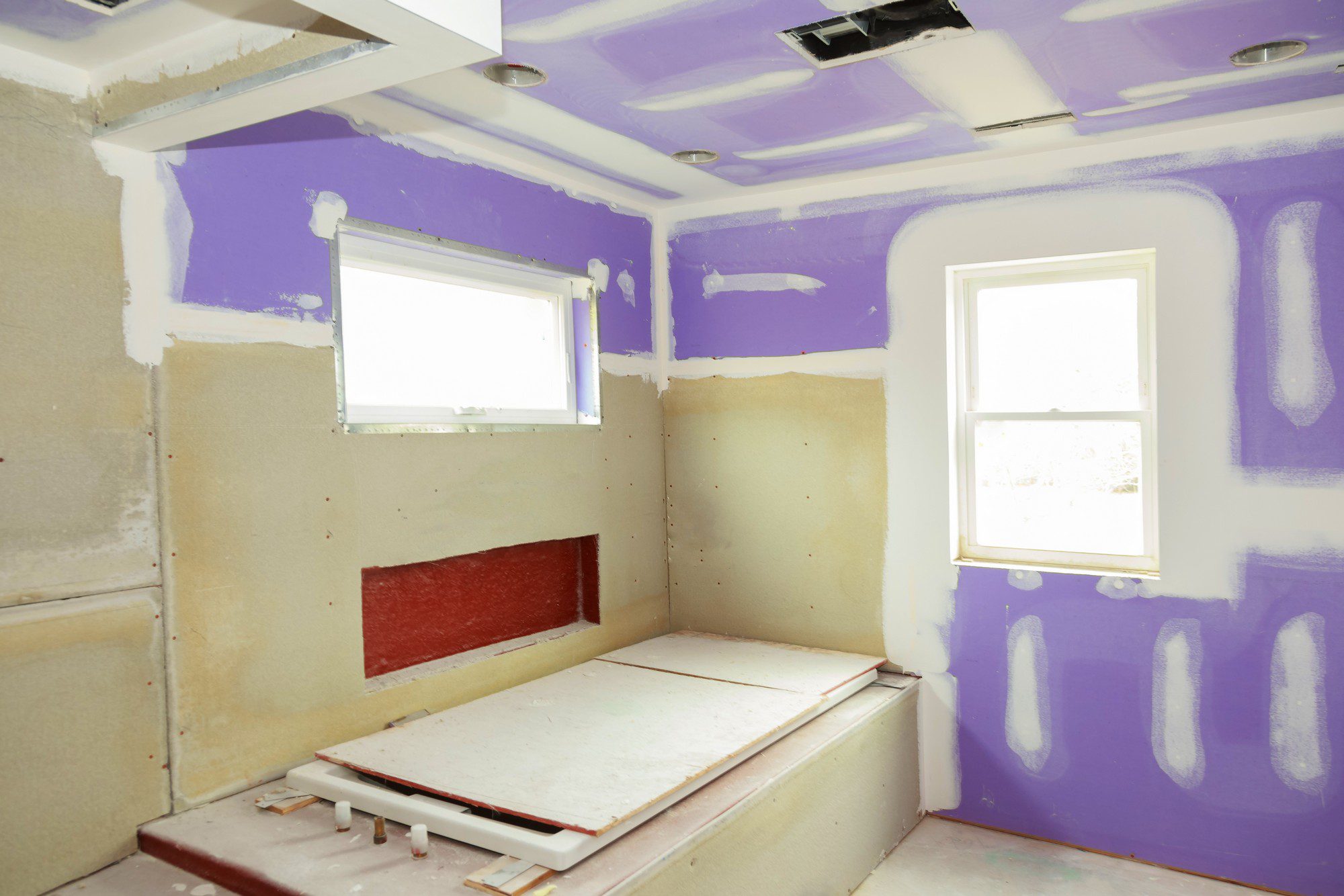 This image depicts an interior space under construction or renovation. The walls and ceiling are insulated with purple foam insulation and partially covered with drywall, indicating that the room is not yet finished. The drywall joints have not been taped or mudded. There are two visible white windows that let in natural light, and various construction materials, such as wood and piping, are scattered around the room. The floor has red-coloured construction material, possibly an underlayer for future flooring, and there are uncovered recessed ceiling lights. The space is seemingly being prepared for further construction or final finishing touches.