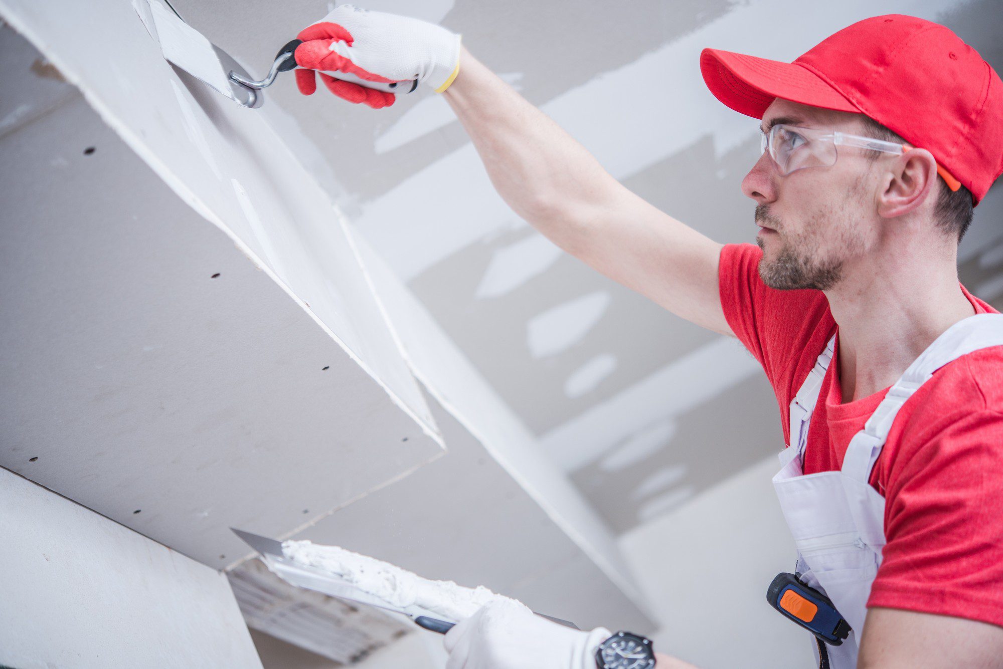 The image shows a man engaging in some construction or renovation work. He is wearing a red cap, safety glasses, a red shirt with a white collar, and white gloves with red detailing. A white tool belt is visible around his waist. The man is using a putty knife or a drywall knife to apply joint compound (commonly referred to as "mud") to what appears to be a seam or joint in drywall. He is smoothing out the compound to ensure a flat and even surface, likely as part of the process to finish the drywall before it is painted or covered with wall treatments. The work is typically done to create a seamless appearance on the wall or ceiling by covering screws and seams between sheets of drywall. The environment suggests an interior setting that is under construction or renovation.