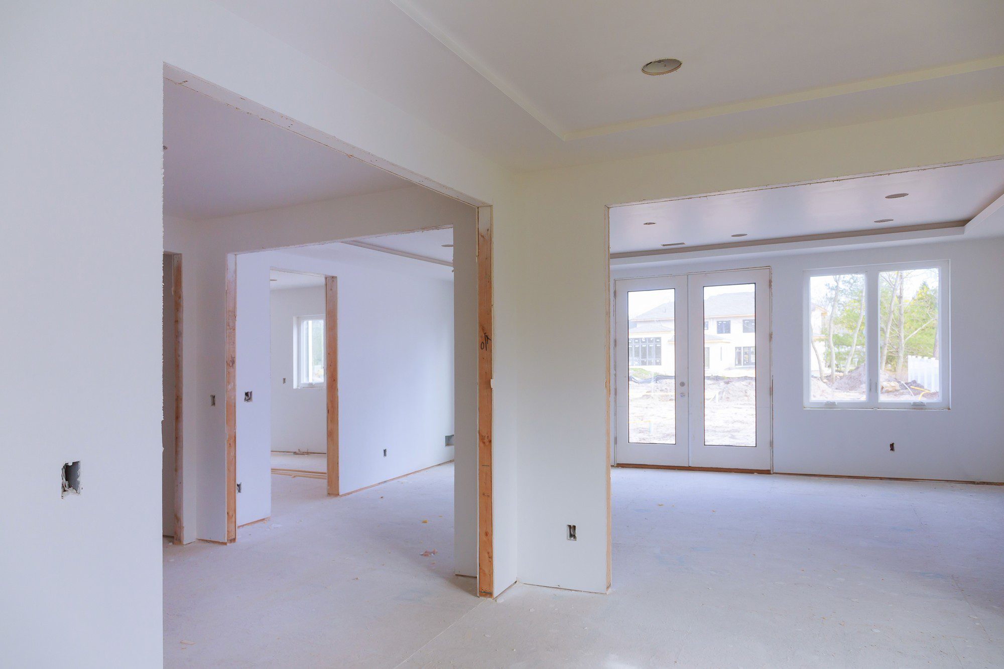 This is an image of a room under construction or renovation. The walls and ceiling have been finished with drywall or plasterboard, and the joints have been taped but not yet fully finished or painted. Electrical outlets and switches have been roughed in, with the outlet boxes visible but without the final covers or faceplates. The floor is subflooring without any flooring material installed. There's a double door with windows that lead to the outside, and a single door in the back wall. The area outside the window appears to be a suburban development, with other houses and construction activity in view.