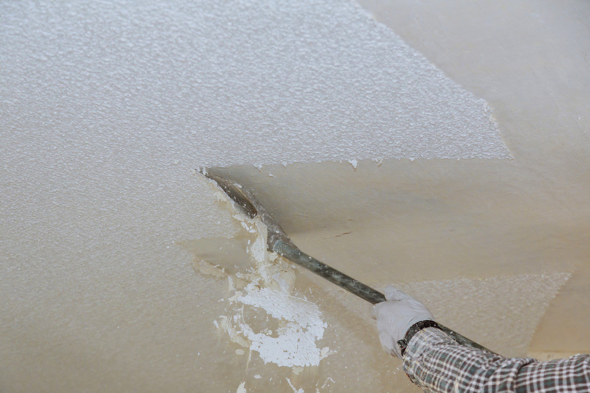 The image shows a person's hand, wearing a white protective glove, using a wide scraper to remove a layer of some white material from a surface. The material that is being removed appears to be flaking off in uneven patches, exposing an underlying layer. The scraper is significantly wide, suggesting that the objective might be to strip a floor, wall, or other large flat surface. The activity could be part of a renovation process, surface preparation for refinishing, or removal of a coating such as paint, plaster, or some other compound. The person seems to be engaging in some manual labour, perhaps related to construction or refurbishment.