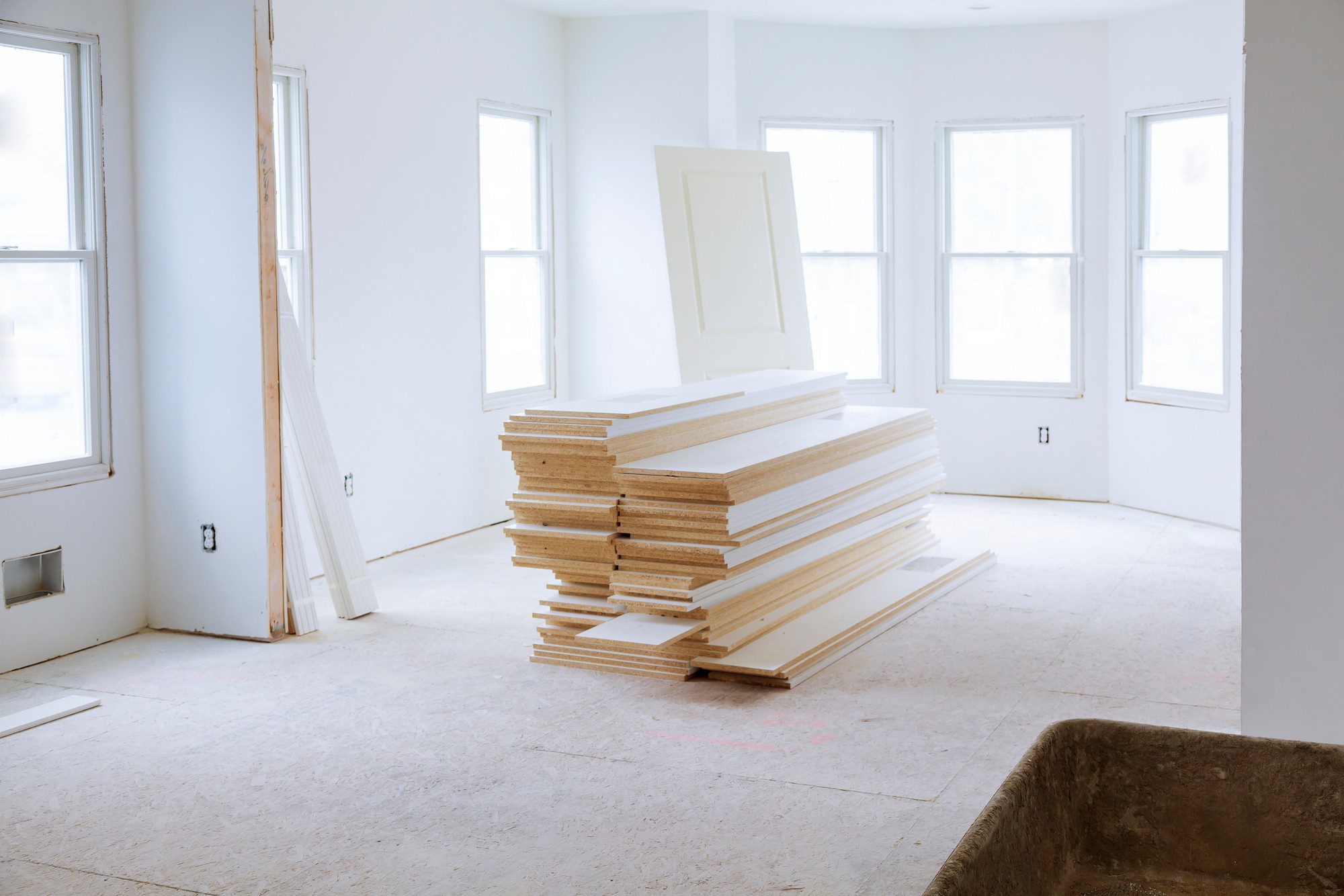 The image depicts an interior space that appears to be under construction or renovation. There's a large stack of unpainted doors or similar rectangular panels in the centre, leaning against the stack is a single panel similar to those in the stack. The space is well-lit with natural light coming through multiple windows. The walls are painted white, and the floor is covered with a protective material, which looks like unpainted particle board or plywood. There are exposed electrical outlets on the walls, and no coverings on the floors or trimming around the room, which is typical for a room that is not yet finished. There's also a pair of wooden boards leaning against the wall on the left side of the photo and a construction cart in the lower right corner.