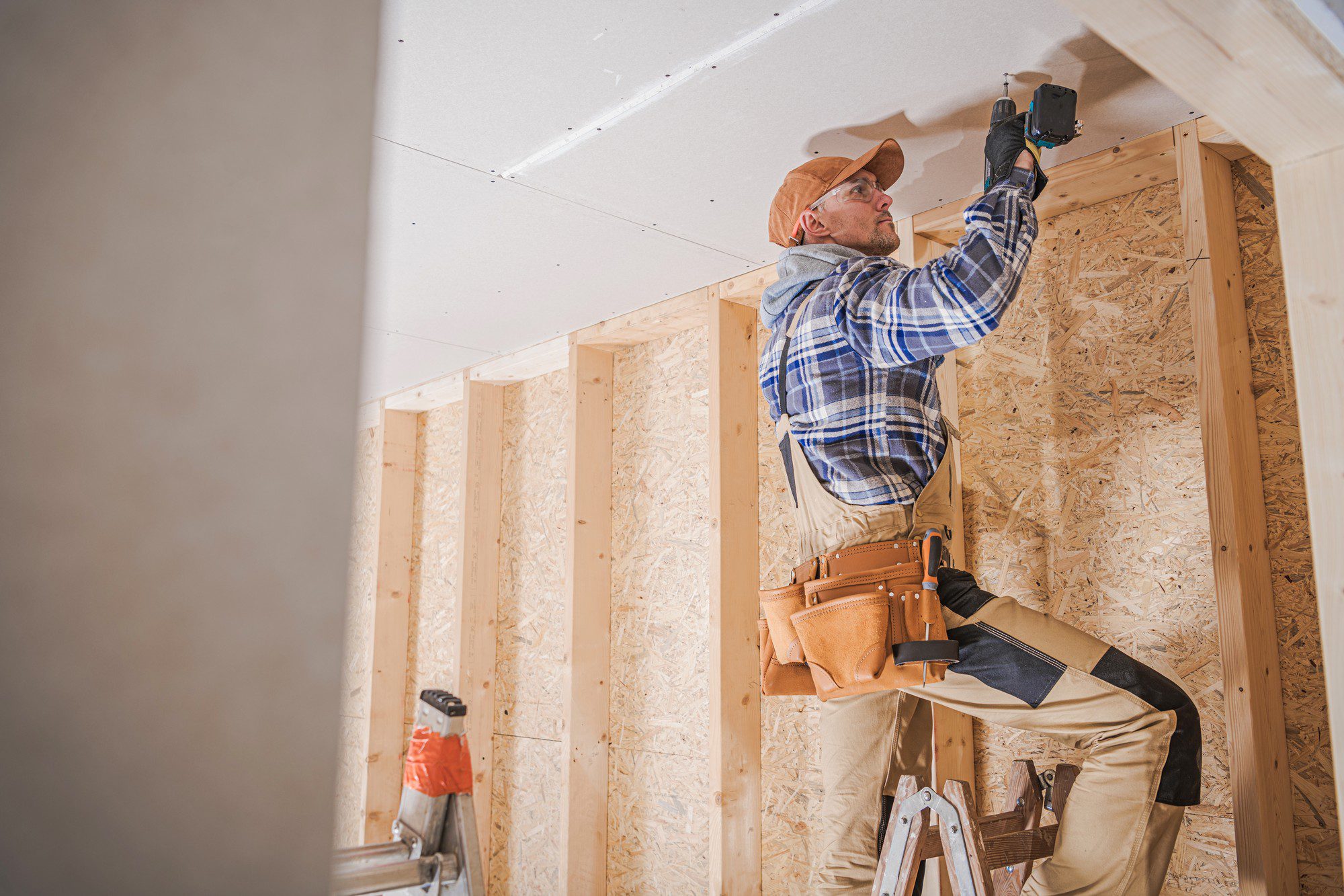 The image shows a construction worker or carpenter who is installing drywall or gypsum board on a ceiling. He is wearing a plaid shirt, khaki pants, a tool belt filled with various tools, and a brown baseball cap. The worker is using a cordless drill or screwdriver to fasten the drywall to what appears to be wooden framing. You can see the structural wood studs and OSB (oriented strand board) sheathing in the background, indicating that the building is in the framing stage. A stepladder is positioned nearby, which he is likely using to reach the ceiling height. The scene is clearly part of an ongoing construction or renovation project.