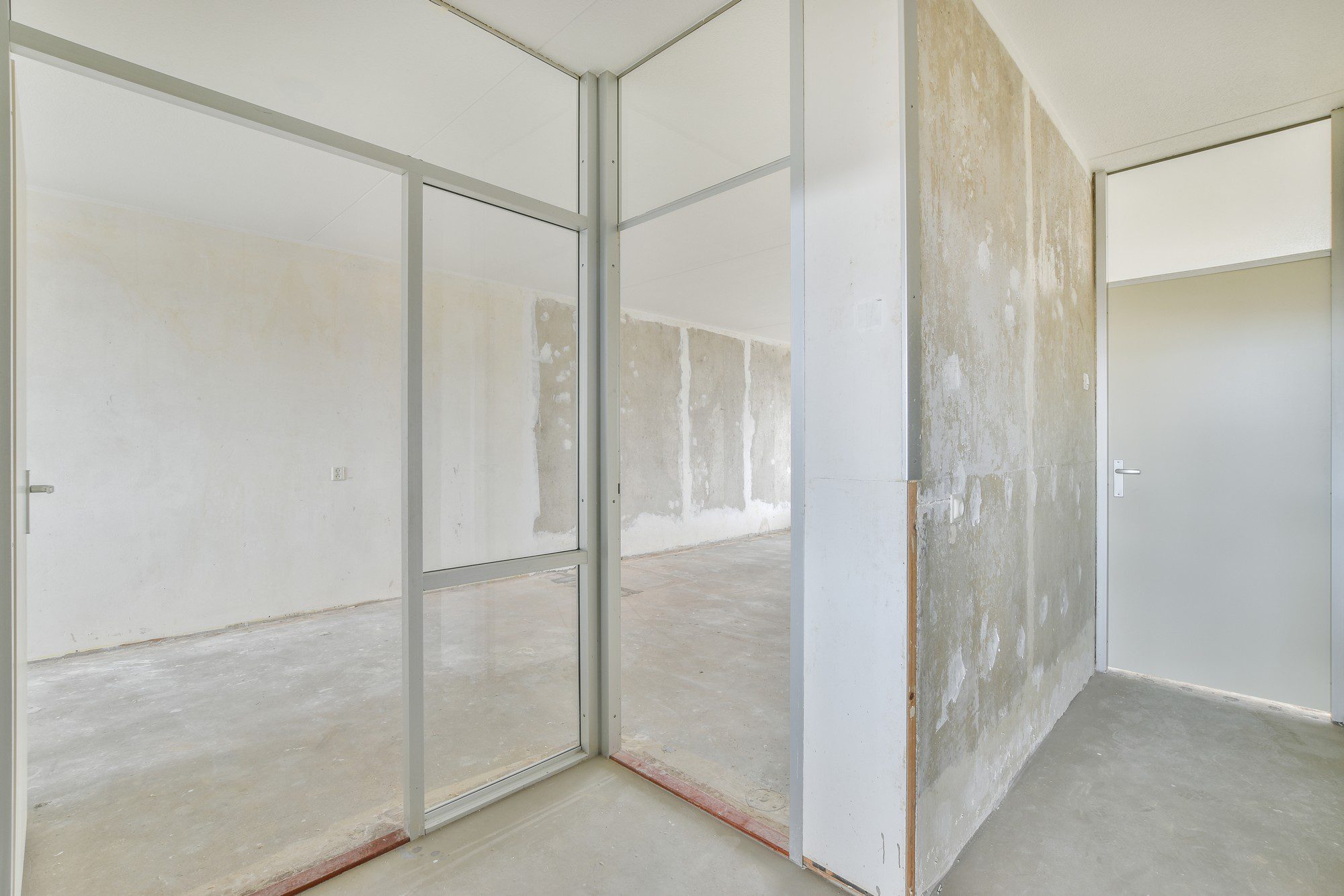 The image shows an empty room undergoing renovation or construction. Visible elements include:
1. A concrete floor that looks unfinished or in need of repair or covering.
2. Walls that show signs of moisture damage or defective plasterwork, suggesting that the space might have been affected by water infiltration or dampness.
3. A glass partition with a metal frame, which appears to separate two sections of the room.
4. There's a closed door on the right-hand side with a modern, simple handle, indicating access to another room or possibly an exit.
5. Electrical outlets on the walls, showing it's a space intended for regular use, probably as an office or similar setting.
6. The ceiling appears to be finished with a white cover, possibly acoustic tiles or drywall, and there's an absence of overhead lighting fixtures.
The space has a general air of being in a transition phase, likely being prepared for renovation or refurbishment.