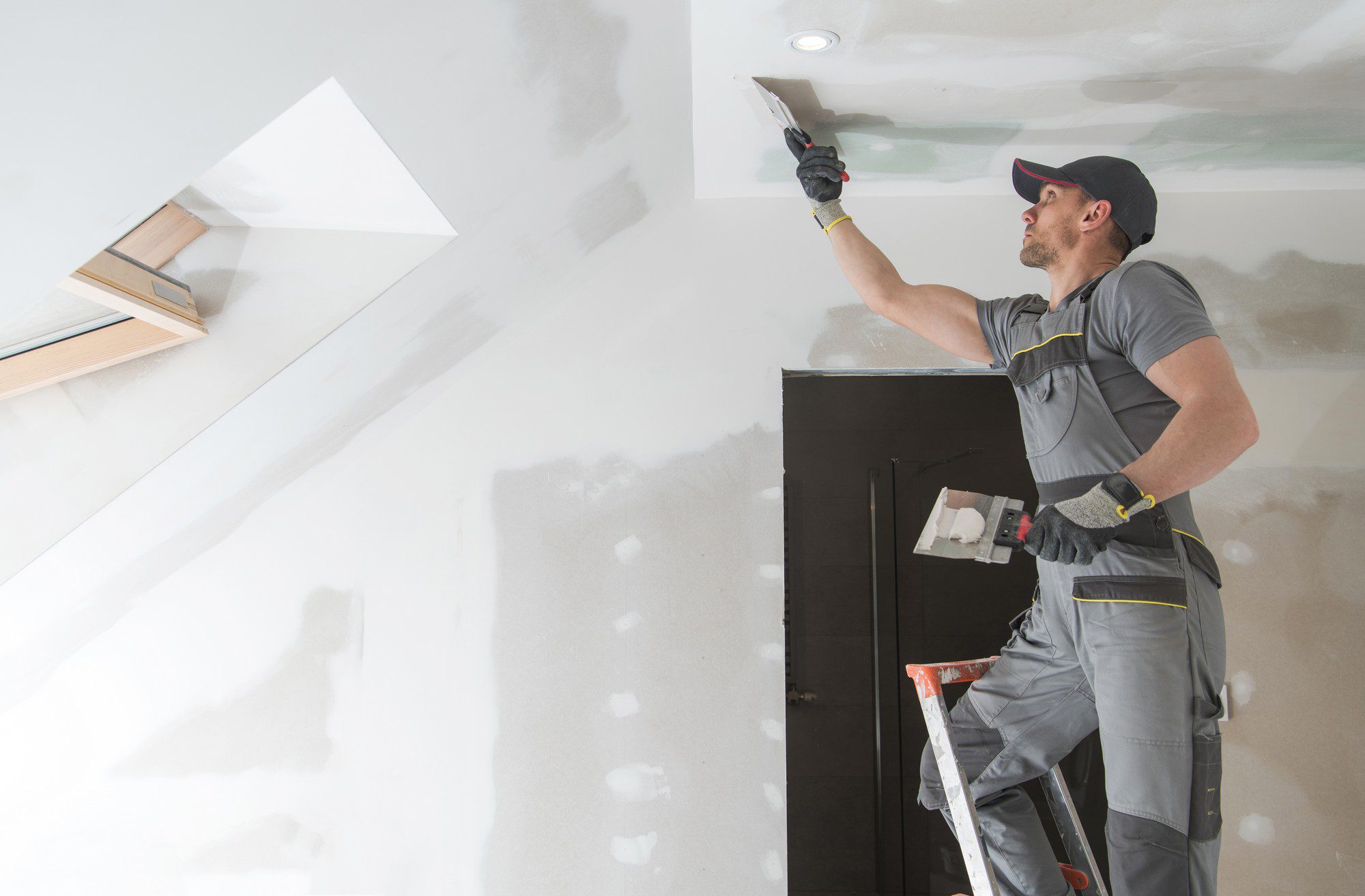 The image shows a person engaged in construction or renovation work, specifically drywall installation or finishing. The individual is dressed in work attire with protective gloves, and is standing on a ladder while using a tool to apply or smooth joint compound (often referred to as "mud") on the drywall ceiling. The ceiling has a skylight installed, and the room appears to be in the middle of construction or remodeling, as evidenced by the unfinished state of the wall with visible joint tape and patches of compound. The person seems focused on ensuring a smooth and even finish for later painting or texturing.