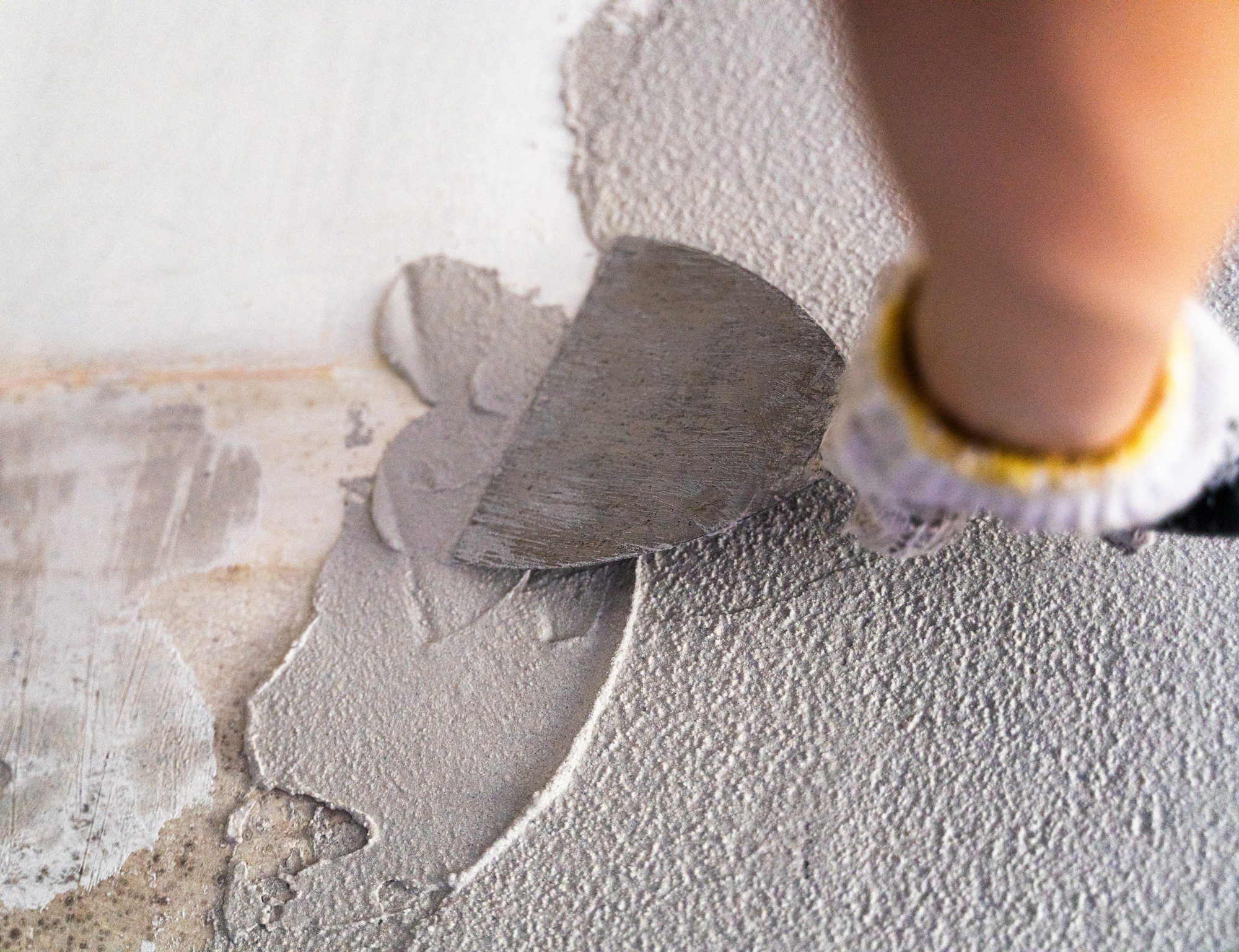 The image shows a person's hand applying a grey substance with a trowel to a wall. The substance appears to be plaster or joint compound, and the person is in the process of smoothing it out over the wall's surface. It appears that they are engaging in some kind of wall repair or finishing work. The wall has a textured surface, and part of it looks like it has been stripped or prepared for the application of this material. The person is wearing a glove, indicating the care taken to avoid direct contact with the material or to keep their hands clean.