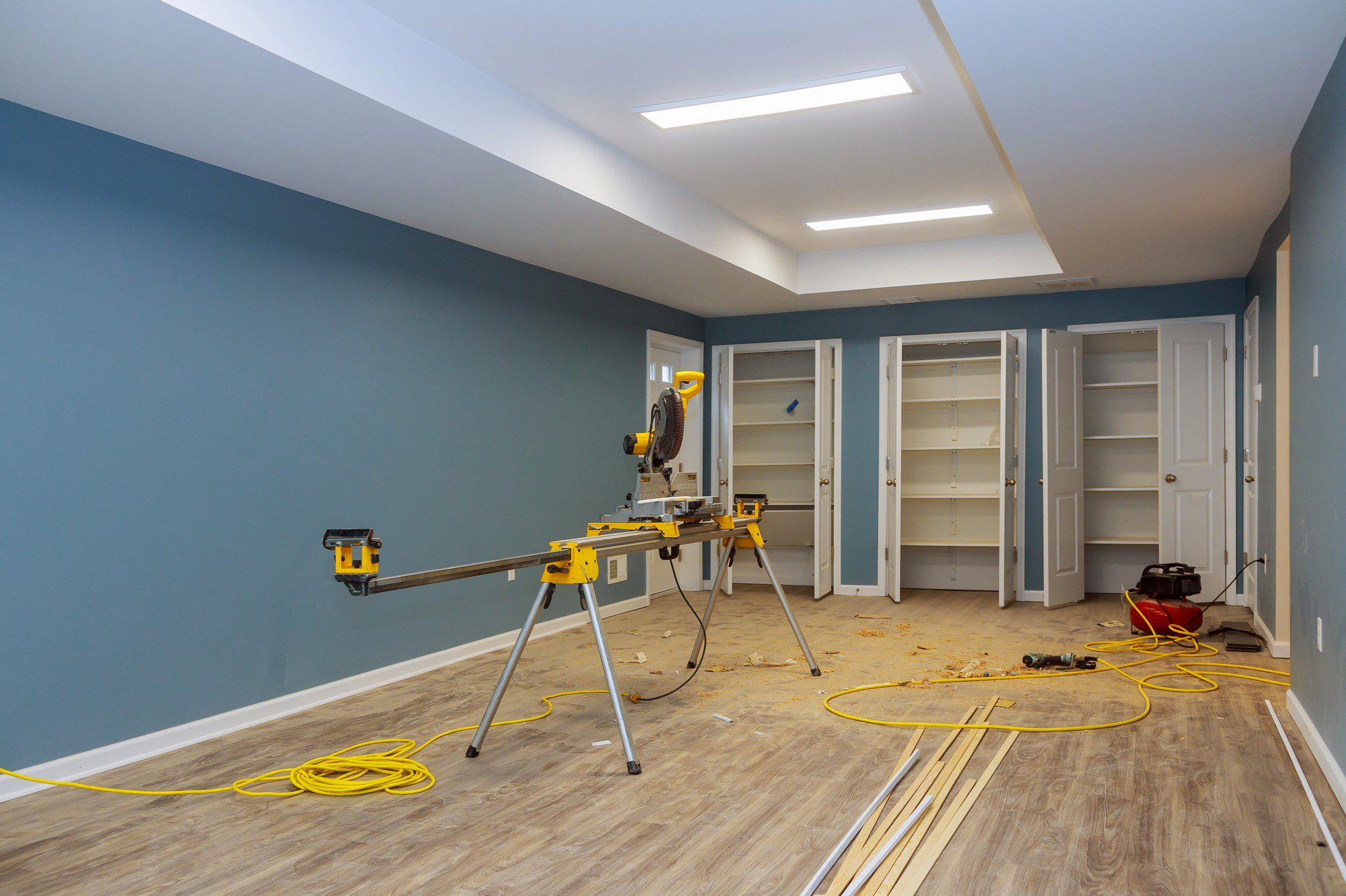 This image shows a room undergoing some form of renovation or construction work. The features visible include:
- **Miter saw**: A power tool used for making precise cuts in wood, often used in trim or finish carpentry. It's set on a stand designed to support longer lengths of wood.
- **Built-in cabinets**: The white cabinets in the background are likely part of a storage or closet system that's being installed or completed.
- **Recessed lighting**: The ceiling has rectangular recessed lighting fixtures installed, providing a modern and streamlined look.
- **Flooring**: There is wood-like flooring that appears to be laminate or vinyl plank given the texture and pattern.
- **Extension cord**: A yellow extension cord is running across the floor, showing that power tools are being used in this space.
- **Debris and materials**: There are wood cuttings and other construction debris on the floor, indicative of ongoing work. Also, there are strips of unfinished wood, likely trim or molding that will be installed.
- **Paint**: The walls are painted in a grey tone with white trim, suggesting that painting has been done recently or is mostly complete.
The room appears to be in a transitional phase, moving towards completion with most of the major installations already in place.