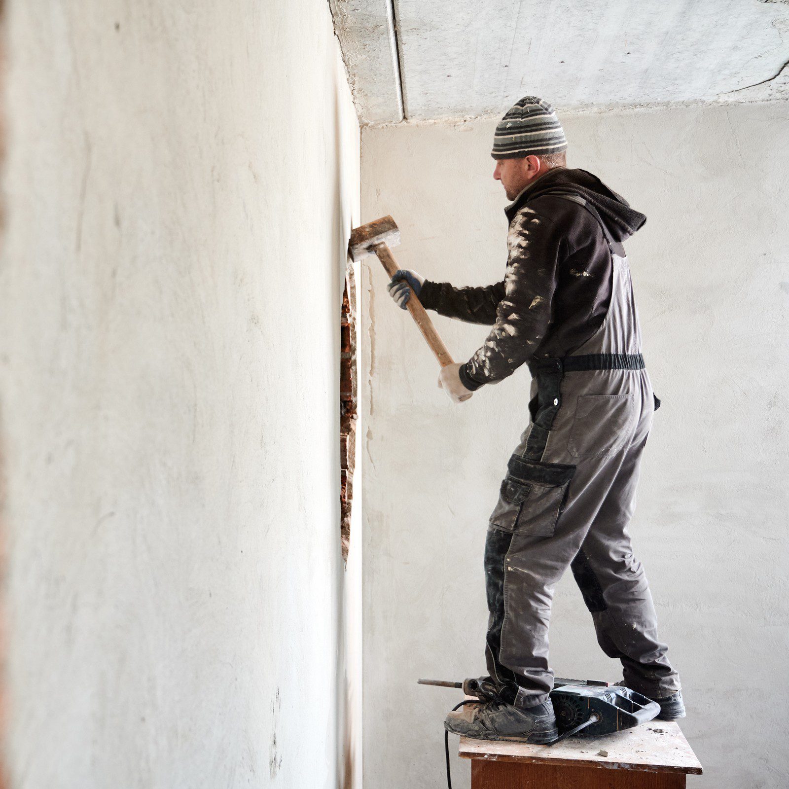 The image shows a person engaged in some kind of manual labour, possibly construction or renovation work. The person is standing on what appears to be a makeshift platform or bench, wielding a hammer and aiming at a vertical surface that might be a wall. They are dressed in work attire, which includes a greyish jumper or jacket, trousers with practical pockets, gloves, and sturdy footwear. The clothes look spattered with white dust, which suggests they might have been working with plaster or other building materials. The scene takes place indoors, and there's a partially exposed edge of a wall or doorway to the right, indicating some demolition or restructuring work is underway. The wall surface looks uneven and could be in the process of being chipped away or smoothed out.