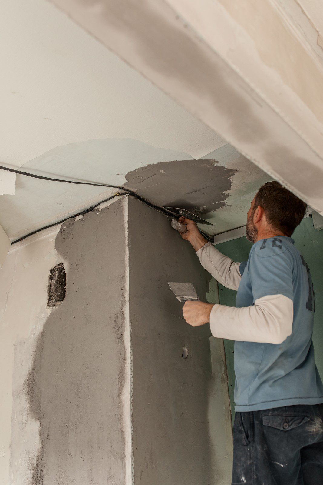 This image shows a person applying plaster to a wall to smooth out the surface or repair damage. The individual is wearing casual work attire and using a trowel to spread the plaster. The wall has areas that are partially covered with a layer of gray plaster with varying levels of smoothness and coverage, indicating the work is in progress. The electrical wiring is visible along where the wall meets the ceiling, suggesting that more work is planned. The setting appears to be an interior space undergoing renovation or repairs.