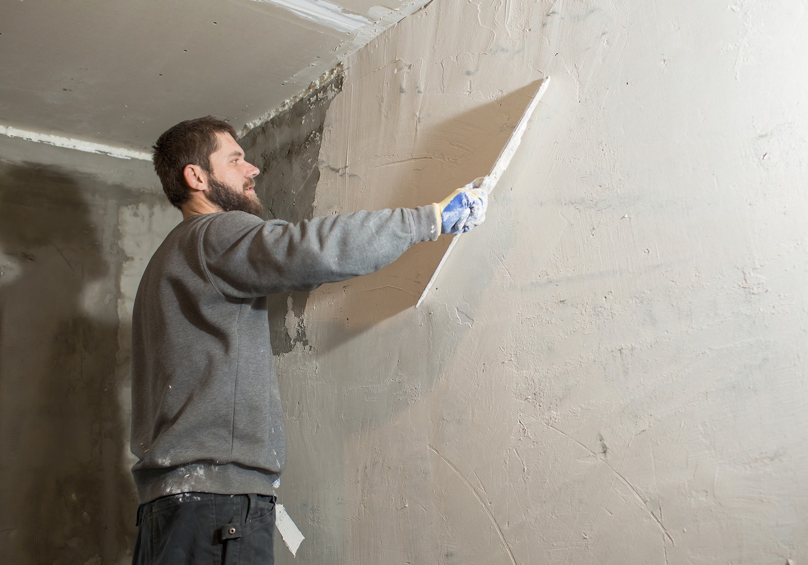 This image shows a person applying plaster to a wall using a large flat trowel or plastering float. The person appears to be in the process of smoothing or leveling the plaster to create a flat, even surface on the wall. They are wearing casual clothing and protective gloves, likely to prevent skin irritation from the plaster material. The wall has areas with a rough layer of plaster, and the individual is working on making it smooth. This is part of the process of wall preparation for either painting or wallpapering after the plaster dries and is sanded down.