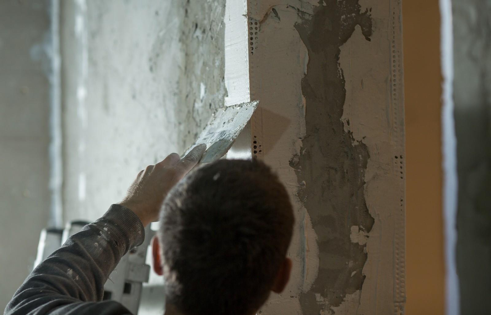 This image shows a person applying a material, likely plaster or joint compound, to a wall with a hand tool. It appears to be during a construction or renovation process, where the walls are being finished or repaired. The person is holding a trowel to smooth the material over the wall surface, which already has some drywall tape and patches applied. The person's focus on the task at hand creates an impression of precision work that is often required in wall finishing.