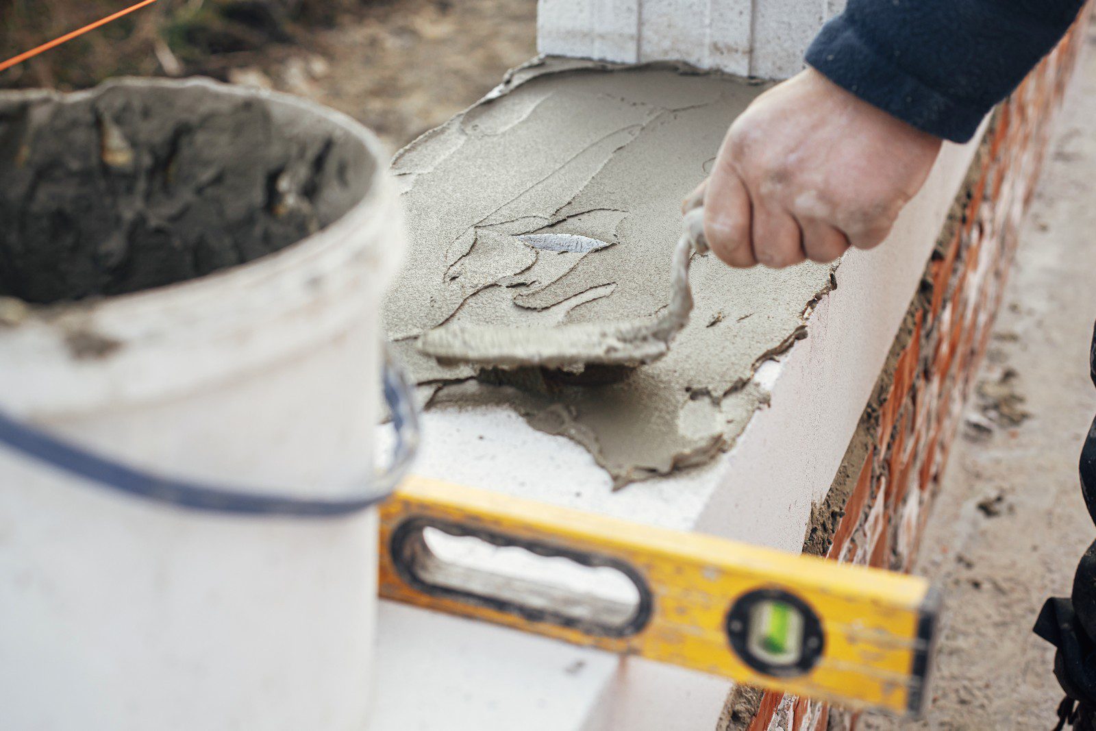 The image shows a person involved in construction work, laying wet cement or mortar on a horizontal surface which appears to be part of a masonry structure. A trowel is being used to spread and smooth the mortar. To the left, there's a bucket that likely contains more cement or mortar mix. In the foreground, a spirit level is placed on the structure, which suggests that the person is ensuring the level and alignment of the work. The presence of brickwork in the background indicates that the work may be related to bricklaying or similar masonry work. It is a clear representation of construction and building practices.