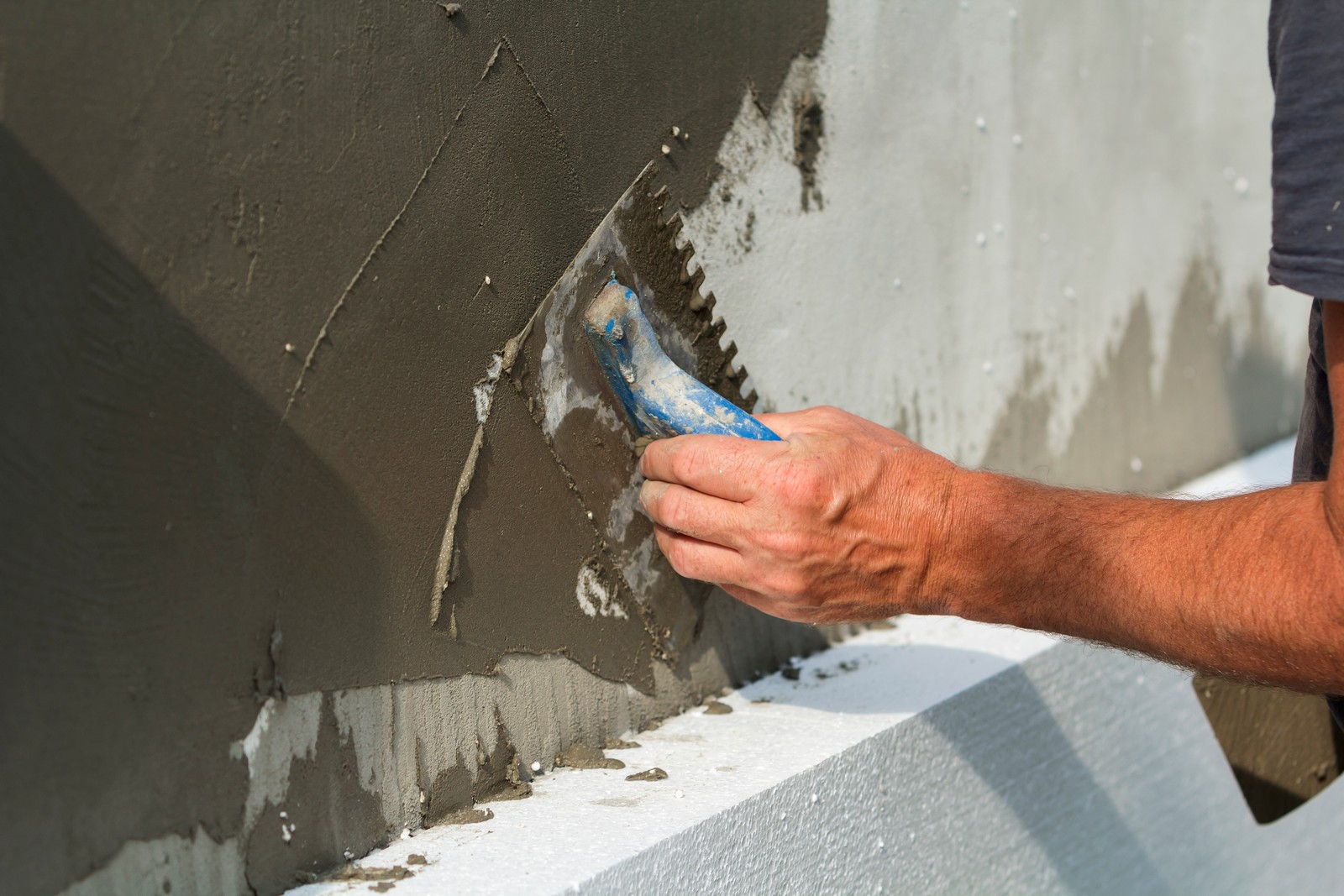 The image shows a person applying a layer of plaster or stucco to a vertical surface, likely a wall. They are using a trowel, which is partially coated with the grey material they are applying. The person's hand is firmly gripping the trowel's handle, and they are spreading the plaster in an upward and sideways motion. The wall section at the top already has a layer of this material, and it seems that the person is working on smoothing and leveling the surface. The person's forearm and hand are visible, and they appear to be wearing a short-sleeved shirt. The focus is on the action of applying the plaster, capturing a moment of construction or renovation work.