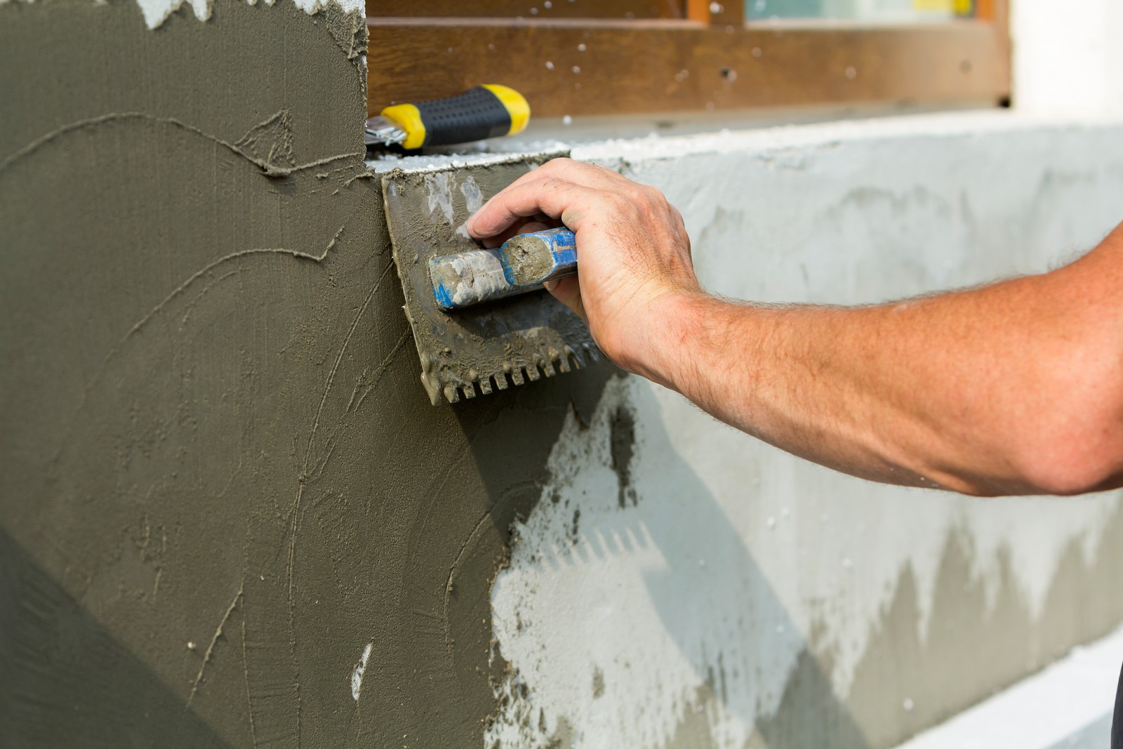 The image shows the hand of a person applying plaster or stucco to a vertical surface with a notched trowel. This process is part of the stages in wall construction or repair, specifically during the application of a base coat or a texture coat which provides an even surface and can also create patterns for aesthetic purposes. There is another trowel or smoothing tool placed on the top ledge above the person's hand, suggesting they are using multiple tools for the job. The person's arm is visible, indicating they are likely in the midst of construction or renovation work.