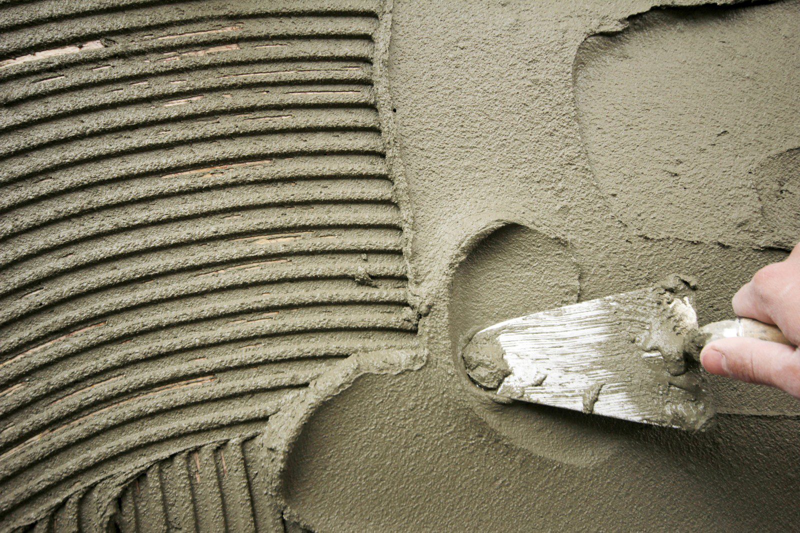 The image depicts a close-up view of a hand holding a trowel and applying wet cement or mortar to a surface. It looks like the person is preparing a wall or floor for tiling since the cement or mortar is being spread in ridges, which is commonly done to ensure tiles adhere properly to the surface. The repetitive lines indicate that a notched trowel has been used to create an even spread with grooves. The photo captures the process of applying adhesive in preparation for laying tiles or similar work in masonry or construction.