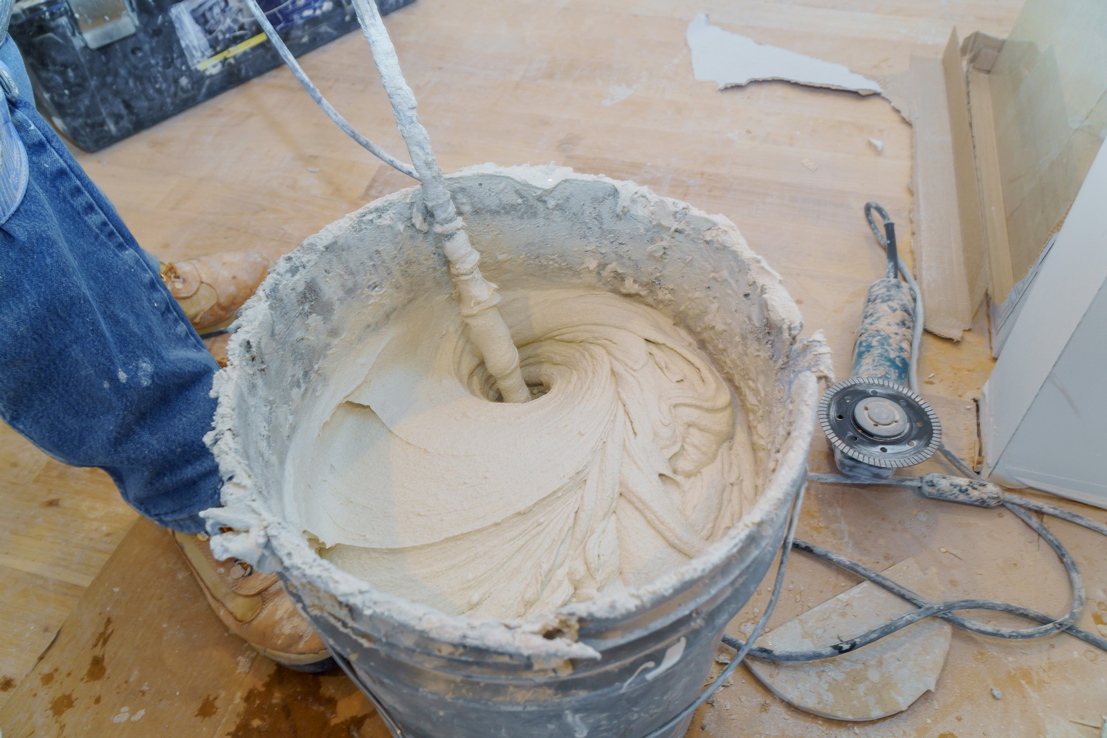 This image features a construction or home renovation scene in which we can see a person's legs wearing blue jeans and brown boots. The person is standing beside a plastic bucket filled with a mixture that appears to be joint compound or plaster. This mixture is being stirred using a mixing paddle attached to a power drill, which has been paused in the action, creating a swirling pattern in the compound. There is also a drywall sanding disc attached to a power tool lying on the ground beside the bucket, indicating that the person is likely involved in drywall installation or finishing work. The floor is covered with protective material, but some drywall dust and debris is visible, suggesting ongoing work.