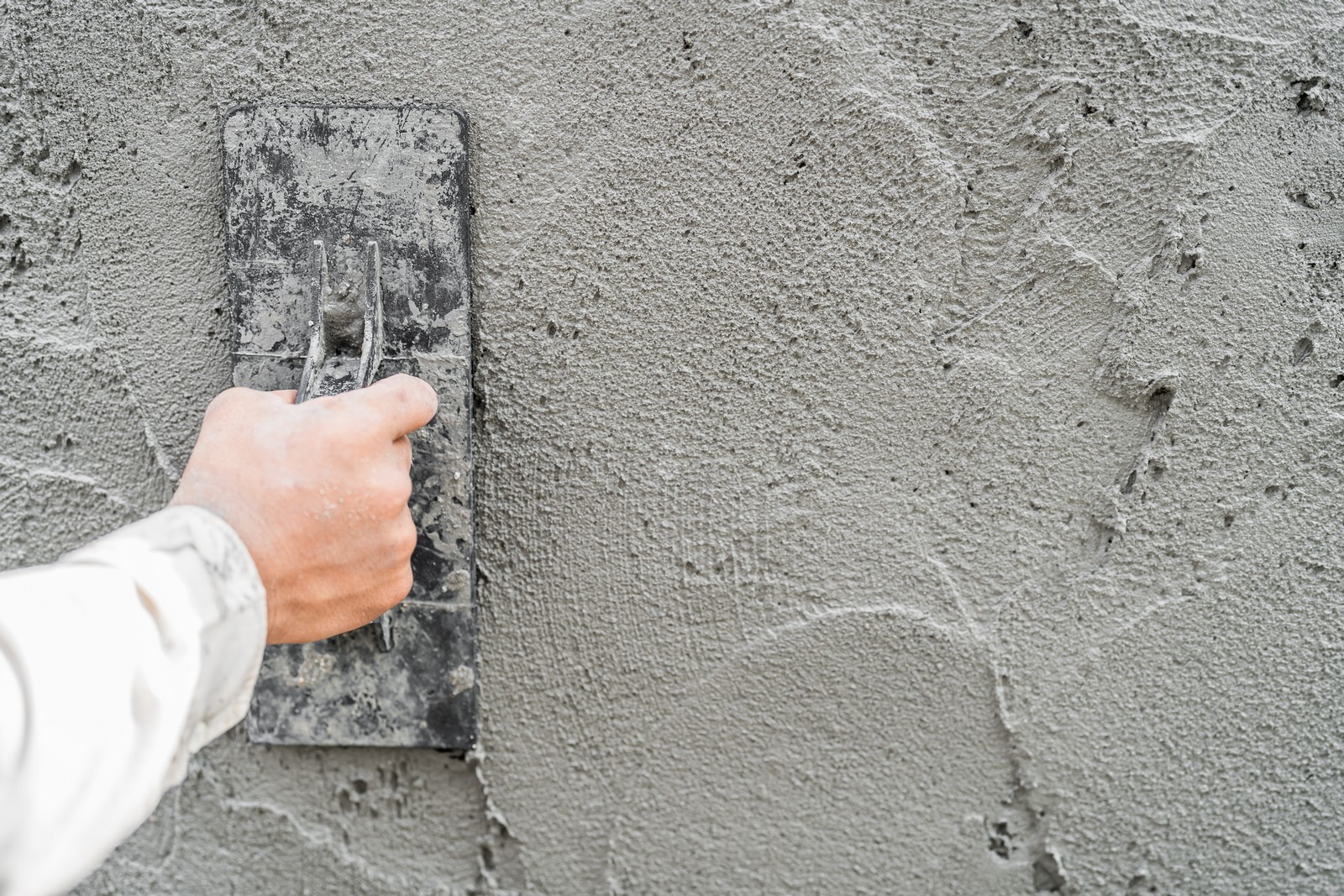 The image shows a person's hand using a plastering trowel to apply or smooth out wet cement or plaster on a wall. The person seems to be in the process of construction or repair work, where the goal is to create a flat and even surface on the wall. The trowel is being held by the handle, and the flat metal surface is in contact with the wet material on the wall. There are also signs of cement or plaster splashes around the area where the work is being done, indicating ongoing work.
