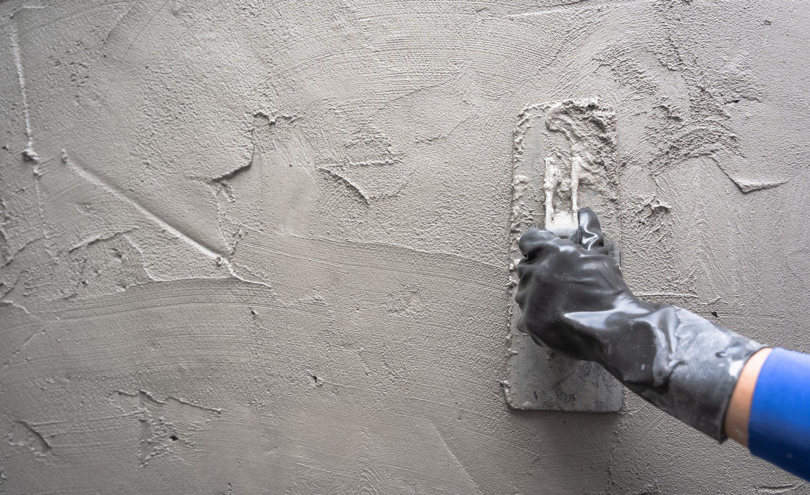 This image shows a person’s hand wearing a black glove and using a trowel to apply or smooth out wet cement or plaster on a wall. The wall is partially covered in the gray material, and the tool is being used to spread and flatten it to create a smooth, even surface. The person appears to be in the process of construction or renovation work, performing tasks related to masonry or plastering.