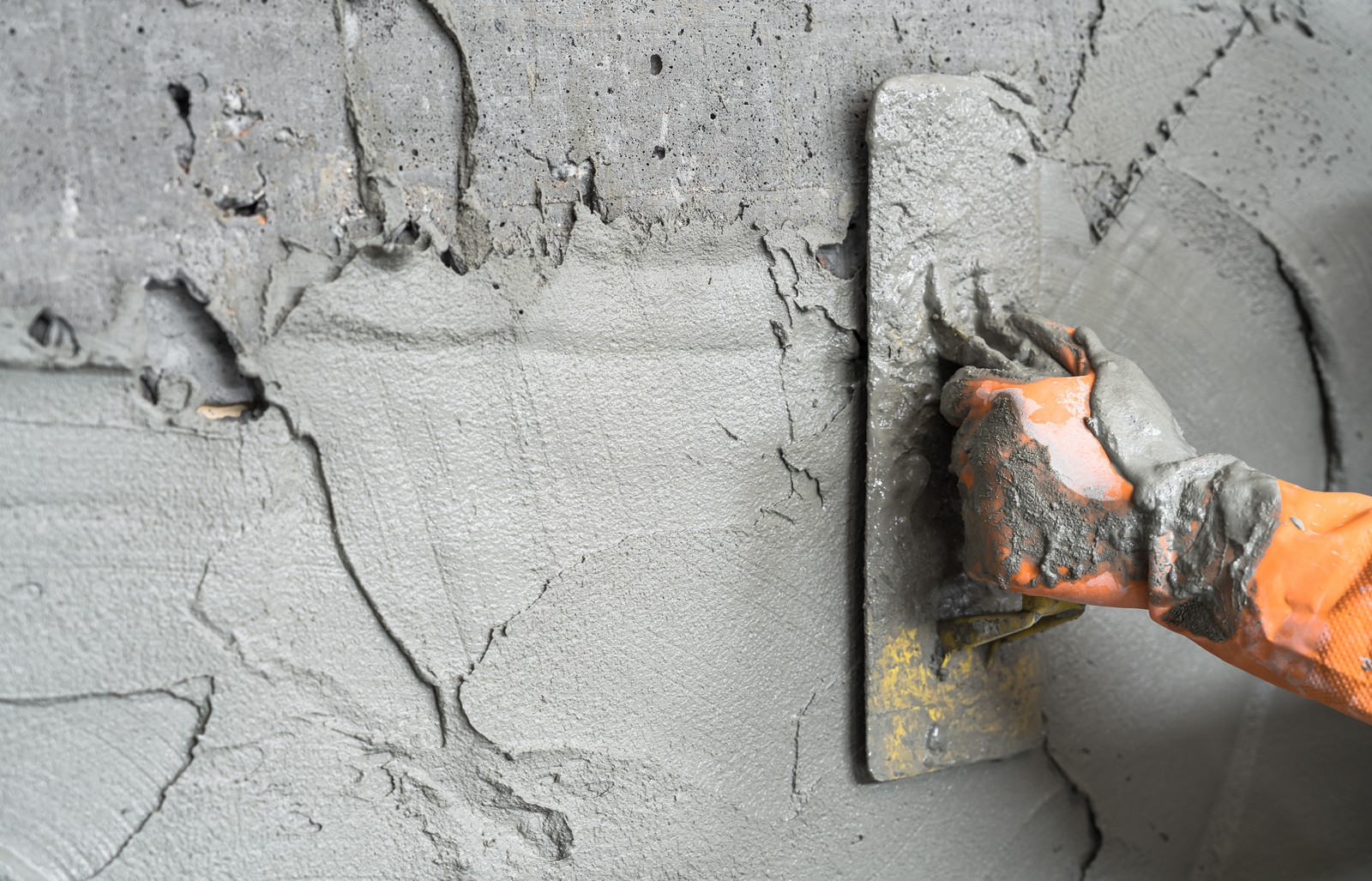 This image depicts a close-up view of a hand in an orange glove using a steel trowel to apply a gray substance, likely wet concrete or mortar, to a vertical surface. The photo shows the hand smoothing the mixture onto the surface, which could be part of a wall or structure being constructed or repaired. Some of the mixture has splattered around, indicating the messy nature of the work.