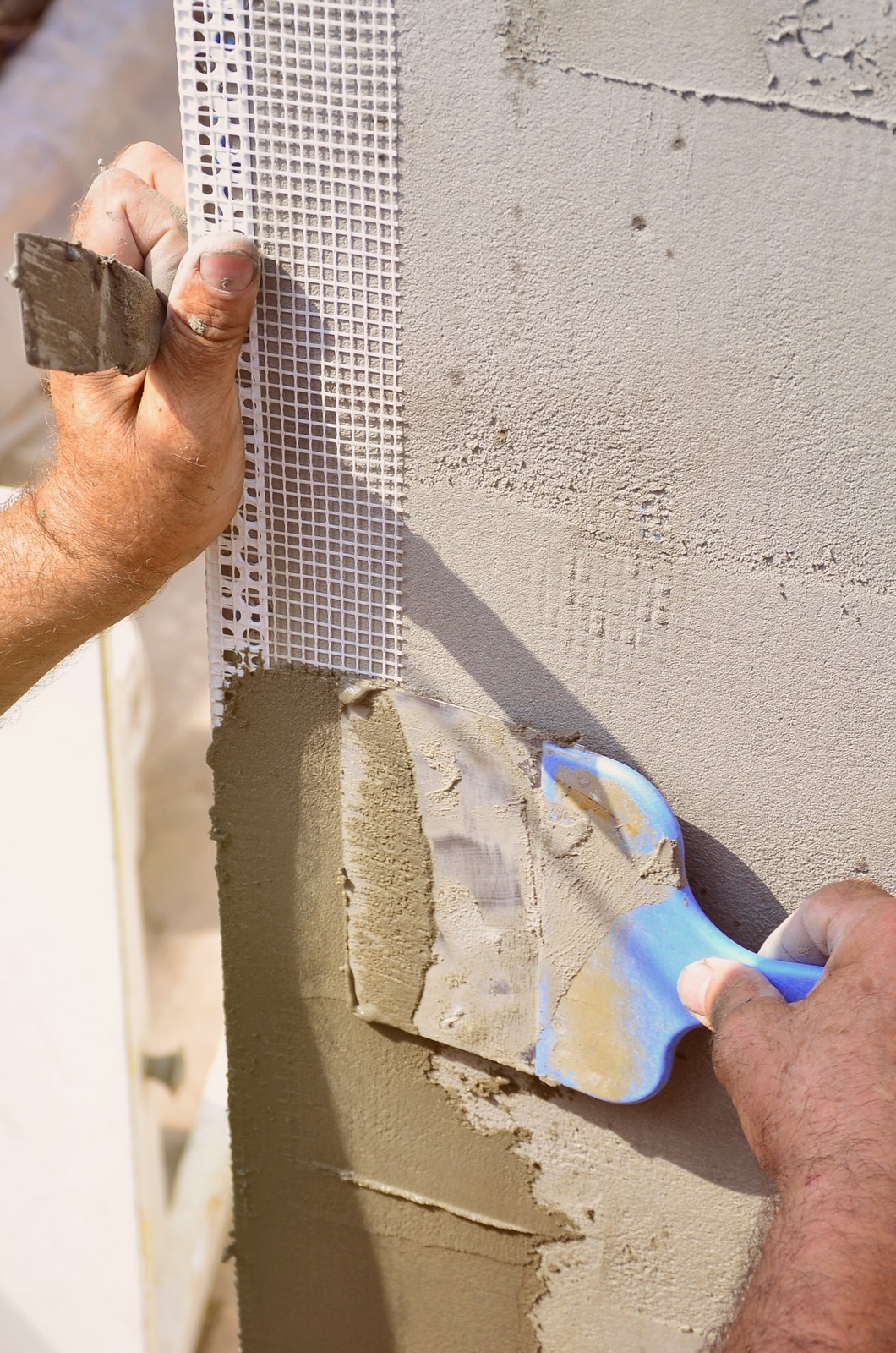 The image shows a close-up of a person's hands applying cement or plaster to a wall as part of a construction or renovation process. The person is using a trowel to spread the material over a mesh, which is commonly used to reinforce the plaster on a wall and to prevent cracking. The individual appears to be skilled in the task, as evidenced by the smooth application and the careful handling of the tools and materials. The picture captures a moment of manual labour typical in the building industry and provides a detailed view of the plastering process.
