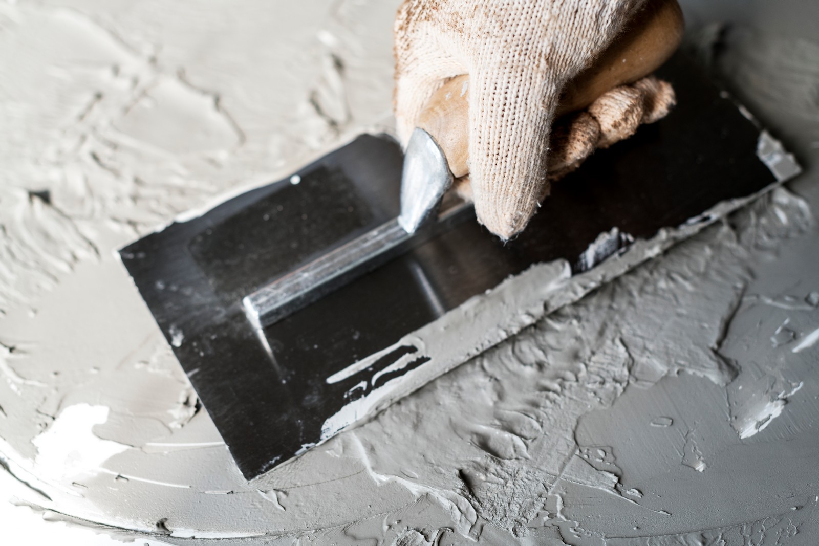 The image shows a close-up of a person's gloved hand holding a metal trowel or putty knife, which is being used to apply and smooth out a gray compound on a surface. The compound appears to be plaster or joint compound, commonly used in building and construction for wall finishing and repair. The glove and tool suggest the person is engaged in a task that requires protection and precision. The background is mostly filled with the same gray material being applied, indicating the work area of the task.