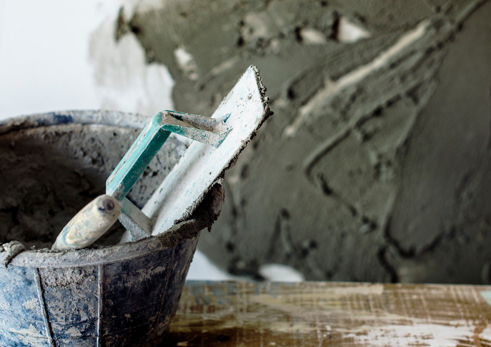 The image shows construction tools and materials. In the foreground, there's a well-used, dirty bucket that seems to contain a mixture, likely cement or plaster, given the context. Resting on the edge of the bucket is a trowel, a tool commonly used for applying and smoothing out these types of mixtures. In the background, we can see a partially finished wall with a smooth, gray coating, which looks like it could be freshly applied plaster or cement. The image gives the impression of an ongoing construction or renovation project where masonry work is being performed.